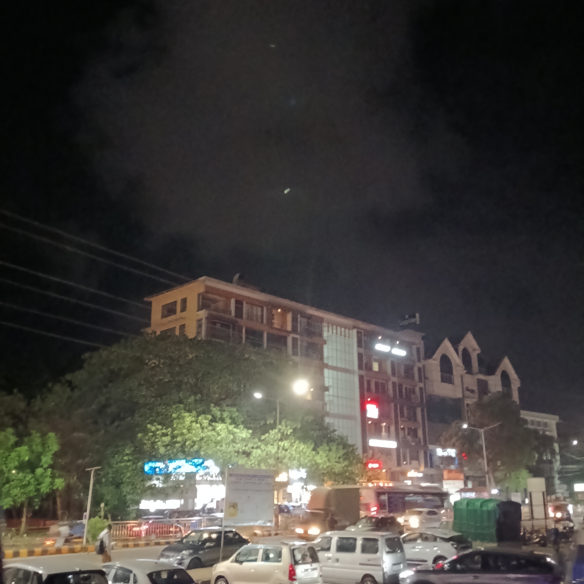 The image shows a street scene in Margao, India, at night. There is a multi-story building with windows and a sign visible. Streetlights illuminate trees and traffic below. Various vehicles, including cars and a van, are parked along the street. A traffic barrier is present, along with pedestrians on the sidewalk. The sky is dark with cloud cover.