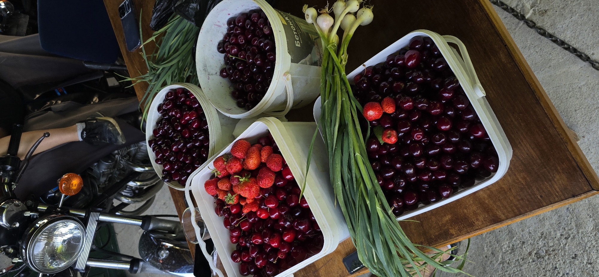 This image depicts a collection of fresh produce, primarily cherries and strawberries, arranged in white plastic buckets on a wooden table. In the background, a motorcycle is partially visible, suggesting an outdoor setting. A person's leg, clad in dark trousers and a grey sock, rests on the motorcycle. Green onions are also present, some of which are bundled together. The scene appears to be a casual display of harvested fruits and vegetables. No specific location in Vratsa, Bulgaria, can be identified from the visual information provided.