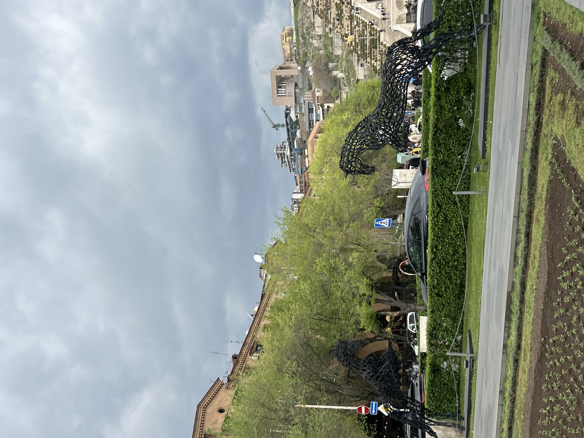 This image, presented vertically, captures a bustling urban scene under an overcast sky. The foreground and mid-ground are dominated by two large, elaborate sculptures of horses or similar creatures, intricately crafted from black, woven metal. These art installations are situated alongside a neatly trimmed green hedge, bordering a paved path or sidewalk. Next to the path, a section of dark soil features small green sprouts.
Behind the hedge, a dark-colored car is partially visible. The landscape includes numerous trees with vibrant light green leaves, suggesting spring or early summer foliage. Various buildings are scattered across the scene; a modern, multi-story structure with a brown facade and large windows is prominent in the mid-ground, while an older, traditionally-styled building with arched openings stands further to the left. A tall construction crane is visible above the buildings, indicating ongoing development.
In the background, a large terraced structure resembling the Yerevan Cascade is evident, with multiple levels and steps. Several small figures of people can be seen on this structure, moving about. The weather is cloudy and overcast, but the lighting suggests it is daytime, likely late morning or afternoon.
Visible text and symbols appear on road signs: a blue square sign with a white pedestrian crossing symbol, a blue circular sign with white arrows (likely a one-way street indicator), and a red circular sign denoting "Do Not Enter." The overall scene depicts a vibrant urban environment in Yerevan, Armenia, characterized by public art, diverse architecture, and typical city activities.