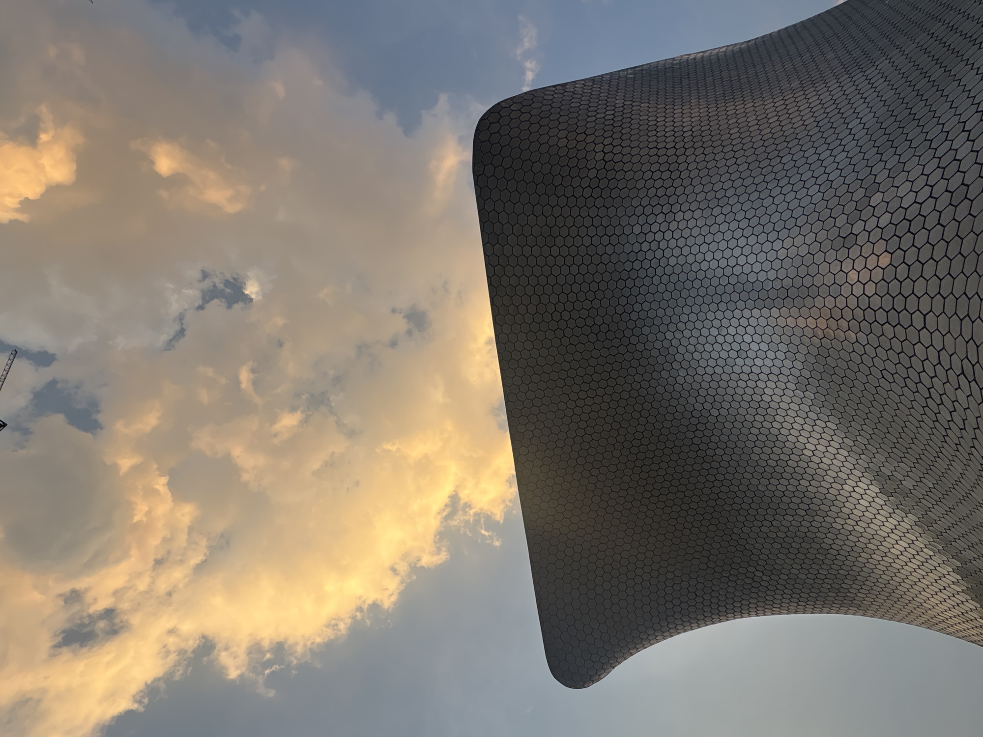 A low-angle shot captures the Museo Soumaya in Mexico City, Mexico, juxtaposed against a cloudy sky. The museum's exterior is covered in hexagonal metal tiles, which reflect the ambient light. The sky is a mix of blue and hues of gold from the setting sun. A construction crane is visible in the distance on the far left, near the corner of the museum.