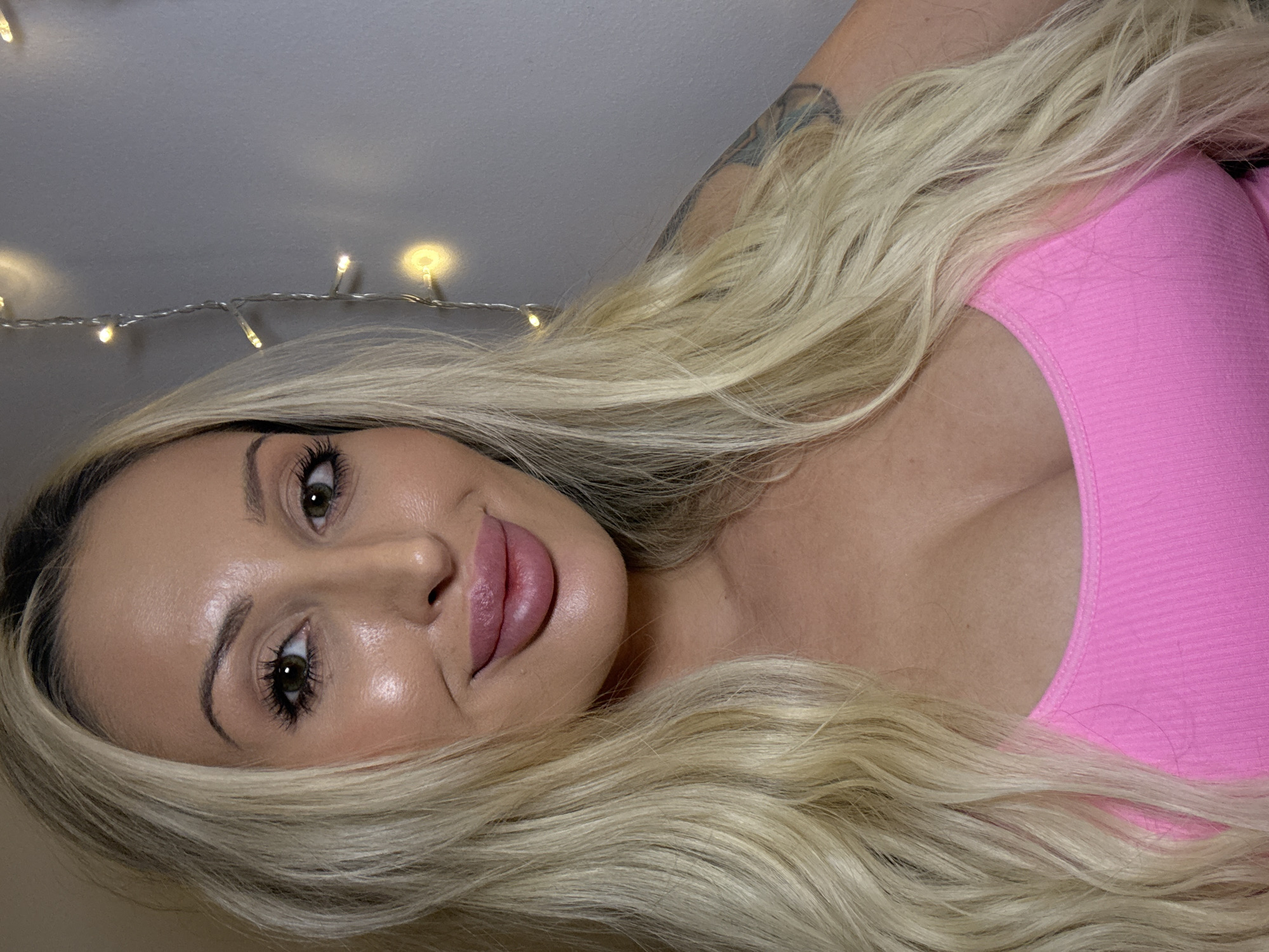 A close-up, head and shoulders shot shows a woman with blonde, wavy hair and fair skin. She is wearing a pink, ribbed tank top and has full lips with pink lipstick. Her eyes are green and she has on mascara and eyeliner. There is a tattoo visible on her upper right arm. The background is a gray wall with a string of fairy lights on the right side of the image. The lighting is warm and casts a soft glow on the subject's face.