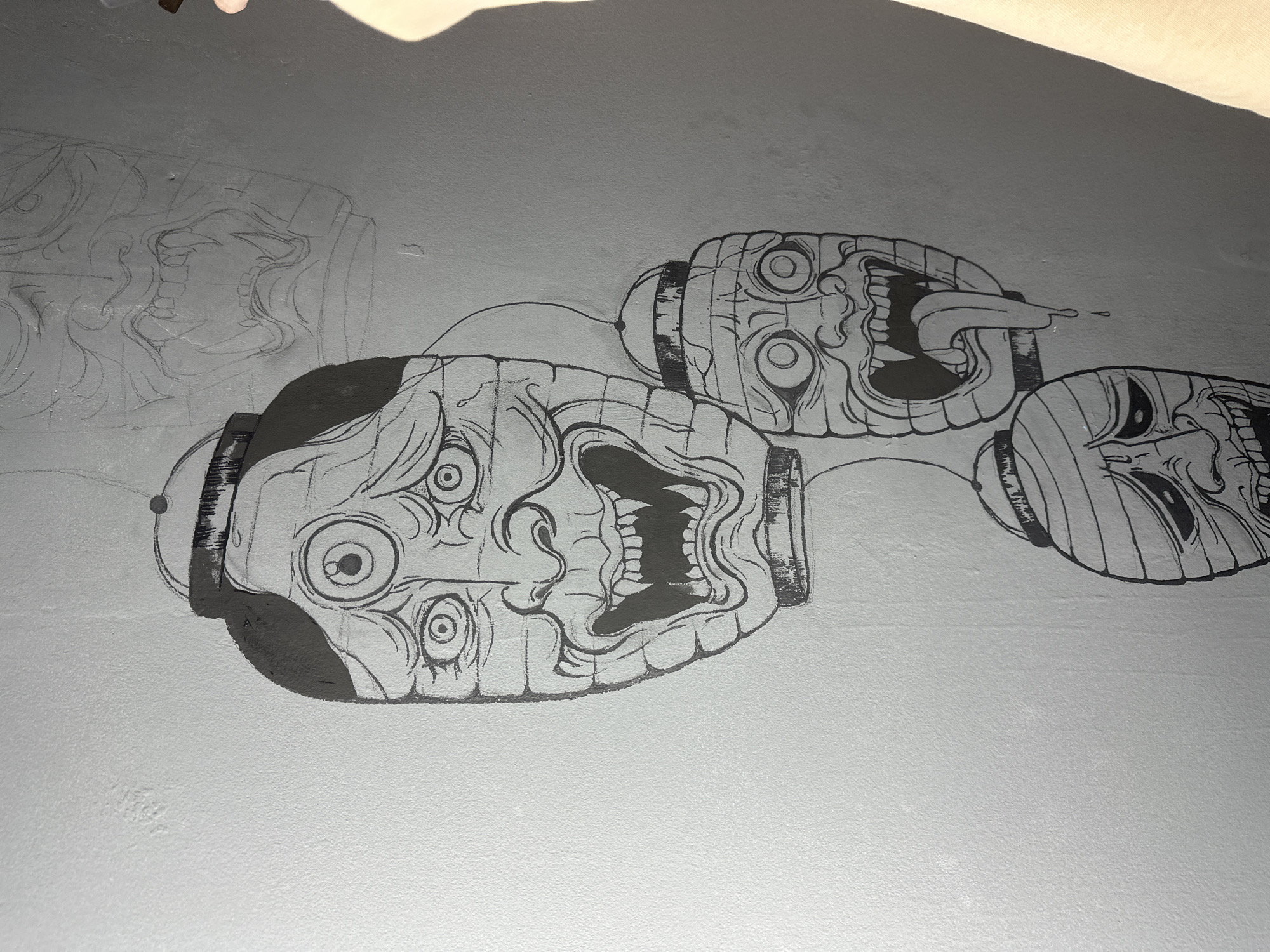 The image shows a close-up of a wall with a partially completed mural in black and white. The mural depicts three stylized Japanese lanterns, each featuring a grotesque or demonic face. The lantern on the lower left is the largest and most detailed, with partially filled black areas. The lantern to its right has a tongue sticking out, with some drool dropping from it. The lantern on the far right has vertical stripes. Fine lines connect the lanterns. There are pencil outlines of another lantern on the left side of the image. The wall is a light gray, with a slightly textured surface. The top of the image has some blurry shadows or a hand partially visible in the frame. The location is unknown, and Saratov, Russia, cannot be confirmed.