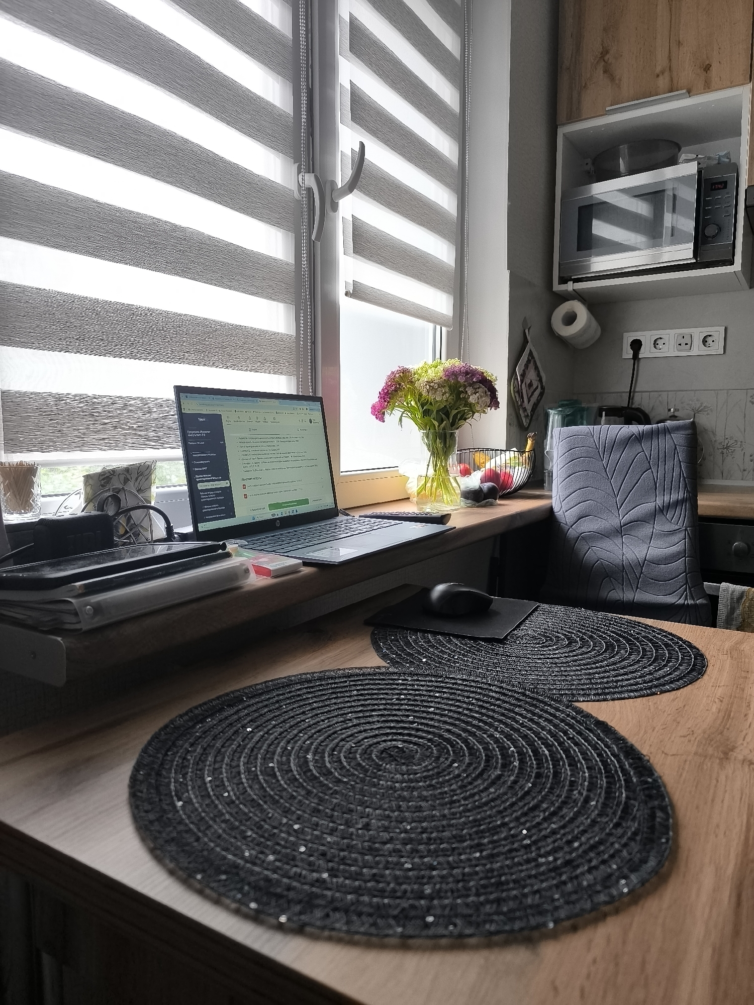 This is a photograph of a home office setup in a kitchen area. The scene is brightly lit, likely by natural light coming through a large window covered by striped roller blinds. In the foreground, a wooden table holds two dark, circular placemats. A laptop is open on the windowsill, displaying a spreadsheet or similar document. To the left of the laptop, there are several notebooks, pens, and what appears to be a tablet. A black computer mouse rests on a black mousepad to the right of the laptop.
Further back on the windowsill, a clear glass vase holds a bouquet of flowers in shades of pink and green. Behind the flowers, a wire basket contains red and yellow fruit. To the right of the basket, a kitchen counter is visible, with a microwave installed in a cabinet above it. A roll of toilet paper hangs on the wall, along with electrical outlets. A grey chair with a leaf-patterned fabric cover is positioned at the counter, partially obscuring the view of the kitchen behind it. The overall impression is of a functional and tidy workspace integrated into a domestic environment. The specific location of Kaliningrad, Russia, is not discernible from the visual elements in the photograph.