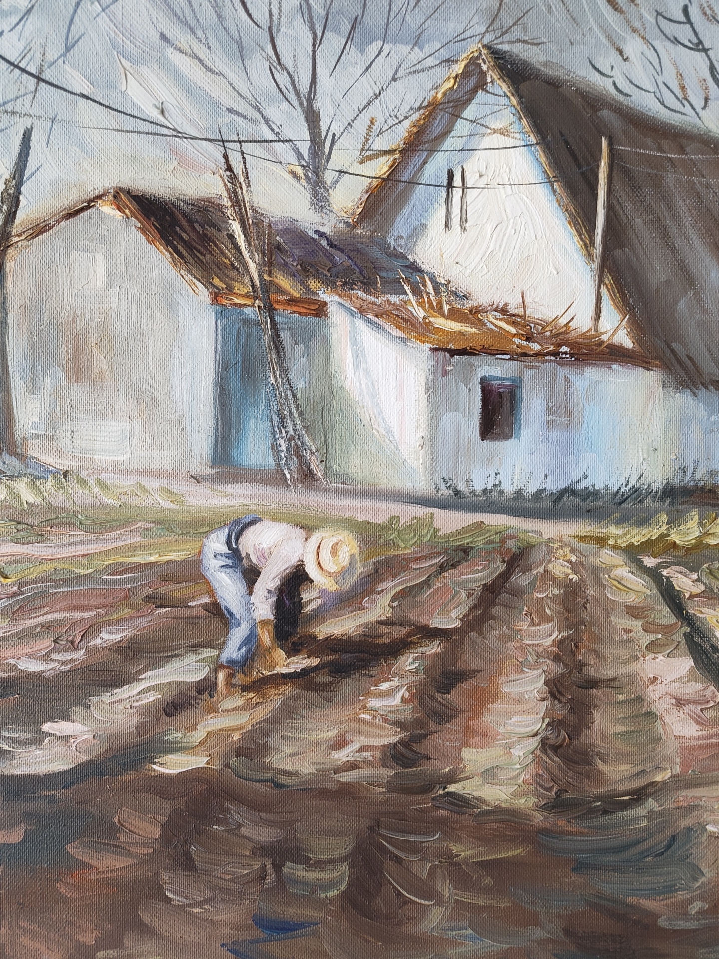 The image is an oil painting depicting a rural scene, possibly in Rotes, Spain, given the traditional architecture. A person, clad in blue overalls, a white shirt, and a straw hat, is bending over in a field. The field is textured with thick brushstrokes of brown and beige paint, suggesting tilled soil. Behind the figure are two whitewashed buildings with thatched roofs. The roofs are primarily brown and grey. The taller building has two vertical rectangular openings, possibly windows, and a small square window lower down. Bare trees are visible in the background against a grey sky, indicating a possibly cold season. The style of the painting is reminiscent of impressionism with its loose brushstrokes and focus on light and color. The location being Rotes, Spain, is only based on speculation, and the painting's actual location is not definitive.