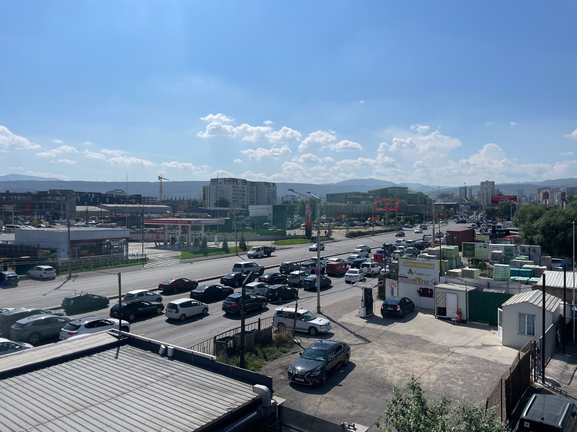 The image depicts a cityscape in Tbilisi, Georgia, under a clear blue sky with scattered white clouds. The foreground is dominated by a corrugated metal roof. A road runs through the mid-ground, filled with a moderate amount of traffic, including cars, SUVs, and a few trucks. On the right side of the road, there is an LPG station with several small buildings, storage containers, and fencing. Buildings and a construction crane are visible in the background, suggesting ongoing development or construction in the area. The scene appears to be a typical urban environment with commercial activity and residential buildings.