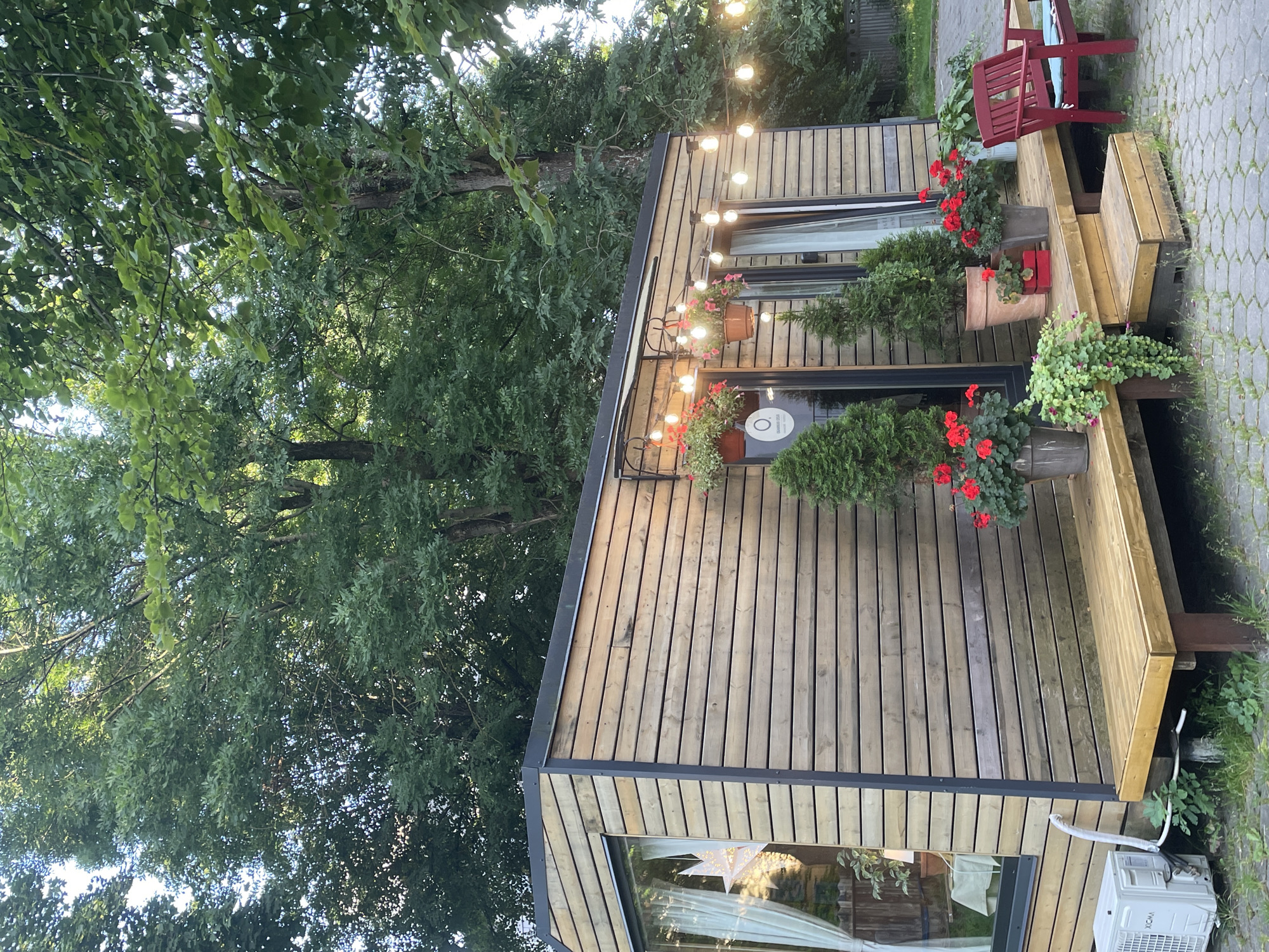 This image captures a scene in Kaliningrad, Russia, depicting a small, modern wooden structure with a deck and outdoor seating. The structure, likely a cabin or tiny house, features horizontal wooden siding, large windows, and a dark-colored roof with an overhang. String lights with incandescent bulbs are draped across the front of the building, illuminating potted plants with red flowers and conical evergreen trees arranged on the deck.
A low wooden deck extends from the front of the structure, with steps leading down to a cobblestone patio. A red wooden chair with blue cushions sits on the patio. To the left of the structure, an outdoor air conditioning unit is visible. The background is dominated by lush green trees, suggesting a park-like or garden setting. The lighting and shadows indicate it is daytime, likely late afternoon or early evening given the warm glow of the string lights. No people are present in the image. The overall impression is one of a cozy, inviting outdoor space, possibly a cafe, guesthouse, or a private retreat.