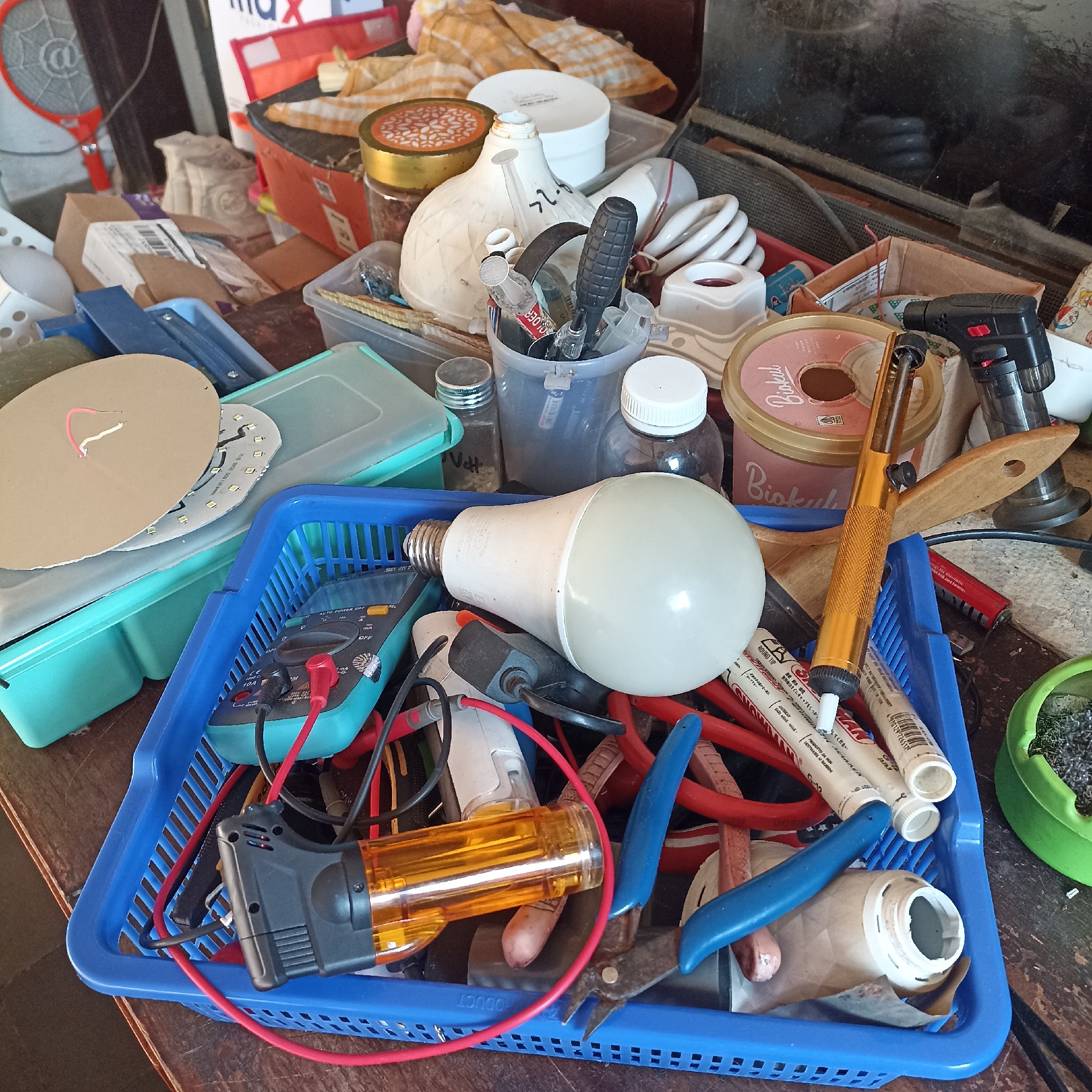 The image depicts a cluttered workbench or storage area in Magelang, Indonesia, filled with various electronic components, tools, and miscellaneous items. A blue plastic basket in the foreground contains a large LED light bulb, a multimeter with red and black probes, a black and orange handheld power tool, red and black wires, and a pair of blue-handled pliers. Other objects visible include a soldering iron, fluorescent light bulbs, jars, containers, and various electrical parts. The overall impression is one of disarray and a collection of items related to electronics repair or DIY projects.