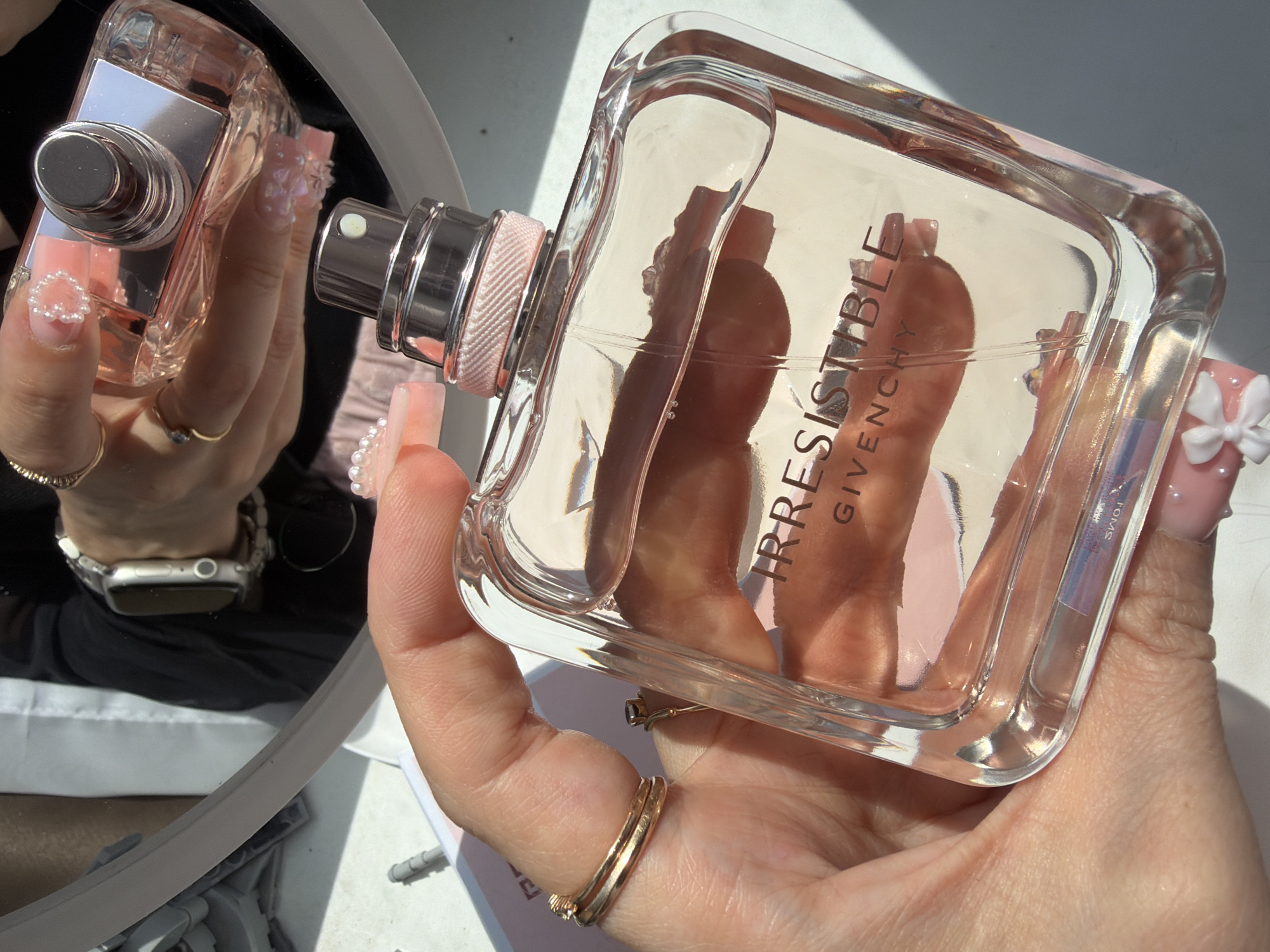 A close-up, high-angle view captures a person's right hand holding an "Irresistible Givenchy" Eau de Parfum bottle in Novosaratovka, Russia. The clear glass bottle, filled with a pale pink liquid, features the brand name embossed on its front and a silver-toned sprayer mechanism with a textured light pink collar. The person's hand displays long, square-shaped nails with a pink base and intricate white 3D floral, bow, and pearl embellishments. Two gold-toned rings adorn the ring finger.
In the upper portion of the image, a reflection in a round or oval mirror shows the person's left hand also holding a similar "Irresistible Givenchy" perfume bottle, held cap-down. This reflected hand exhibits similar pink manicured nails, decorated with white 3D pearl borders and individual pearls. The reflected hand wears gold-toned rings on the index and ring fingers, a silver Apple Watch with a light-colored band, and a beaded bracelet on the wrist. The reflection also reveals part of the person's attire: a black long-sleeved top with a sheer overlay and a glimpse of a light pink garment or background below. The setting appears to be a brightly lit interior, possibly a white table or surface in the foreground, with strong, natural light casting distinct shadows and highlights on the glass and metal surfaces of the perfume bottles. A small, light grey cylindrical object lies on the white surface to the left of the foreground hand.