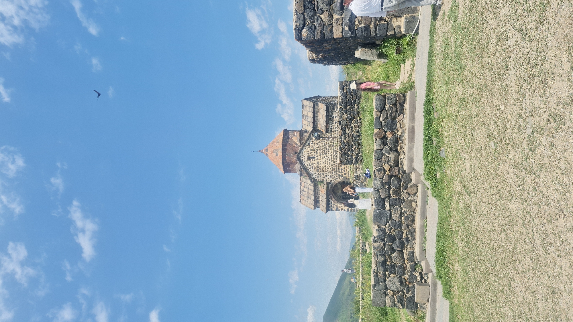 This is a vertical, eye-level shot of a historic Armenian church and its surroundings under a bright blue sky with scattered clouds. The church, built of dark, rough-hewn stones with a distinctive red-tiled conical roof, is the central focus. In front of the church, a low, dark stone wall encloses a grassy area. Several people are visible: two women in white dresses and a person with dark hair and a light-colored top are gathered near the entrance of the church. To the right of the church, another low stone wall is visible, with a young girl in a pink dress standing near a pathway. In the distance, rolling green hills are visible under the expansive sky, and a bird is flying in the upper right portion of the frame. The foreground is a patch of dry grass and dirt. The scene appears to be a tourist site, likely Sevanavank monastery on the peninsula of Lake Sevan in the city of Sevan, Armenia, given the architectural style and landscape.