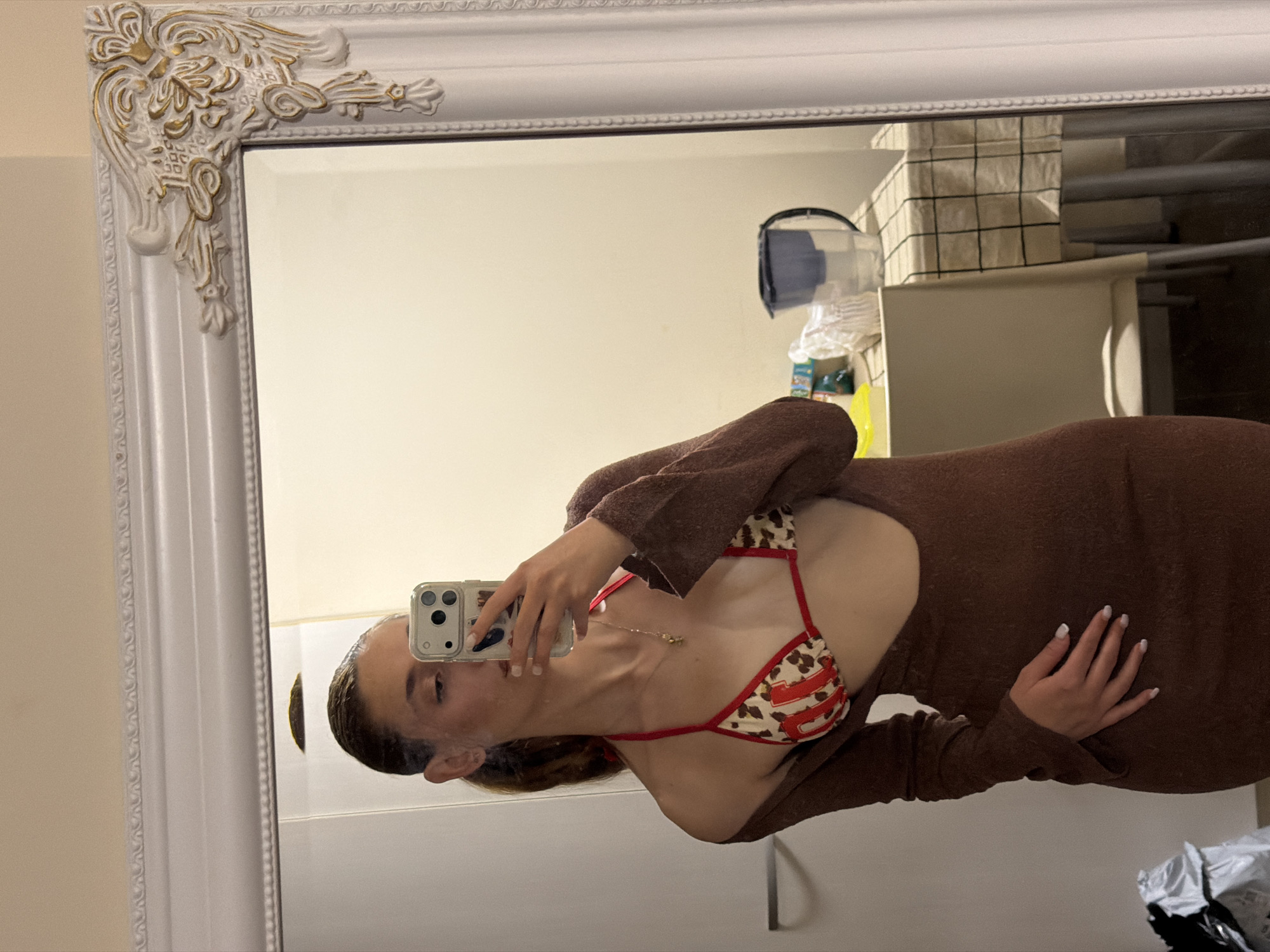 A person is taking a selfie in a mirror. The mirror has a decorative white frame. The person is wearing a brown long-sleeved garment and a bikini top with a red trim and a pattern of beige, brown, and red. A gold necklace is visible around the person's neck. The background shows a white wall and various items, including a vacuum cleaner, cleaning supplies, and a tiled wall. The location is not possible to determine with the objects given.