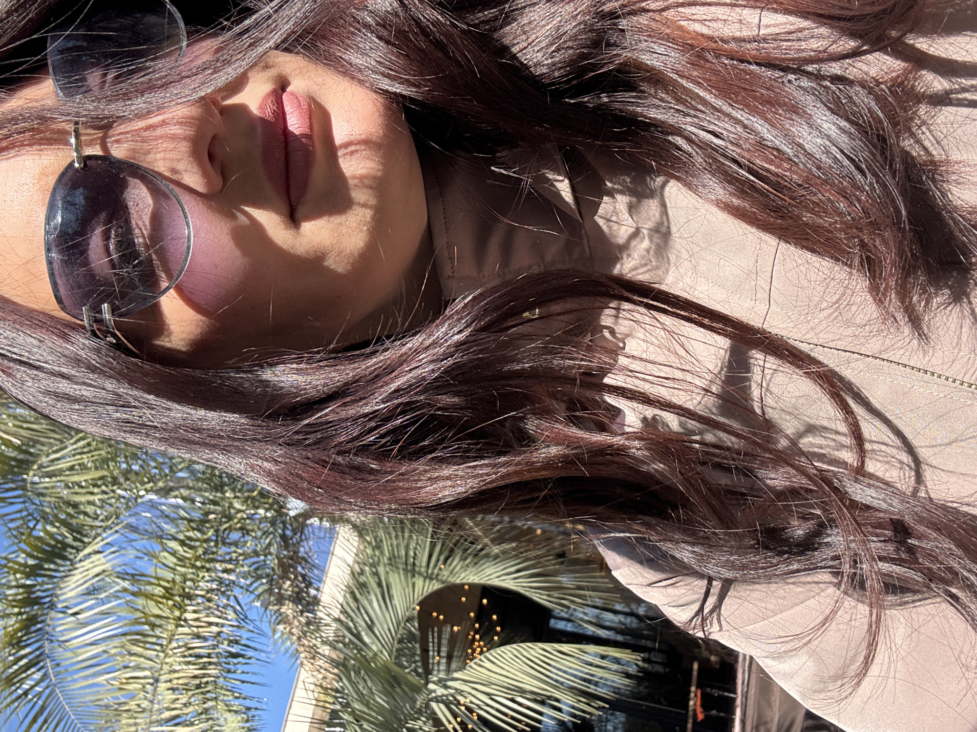 The image shows a person wearing sunglasses and light pink lipstick. Their dark brown hair is visible, and they appear to be wearing a light-colored jacket. In the background, there is a building and palm trees, suggesting a tropical or subtropical environment. The scene is likely located in Sochi, Russia, given the presence of palm trees and the architecture visible in the background.