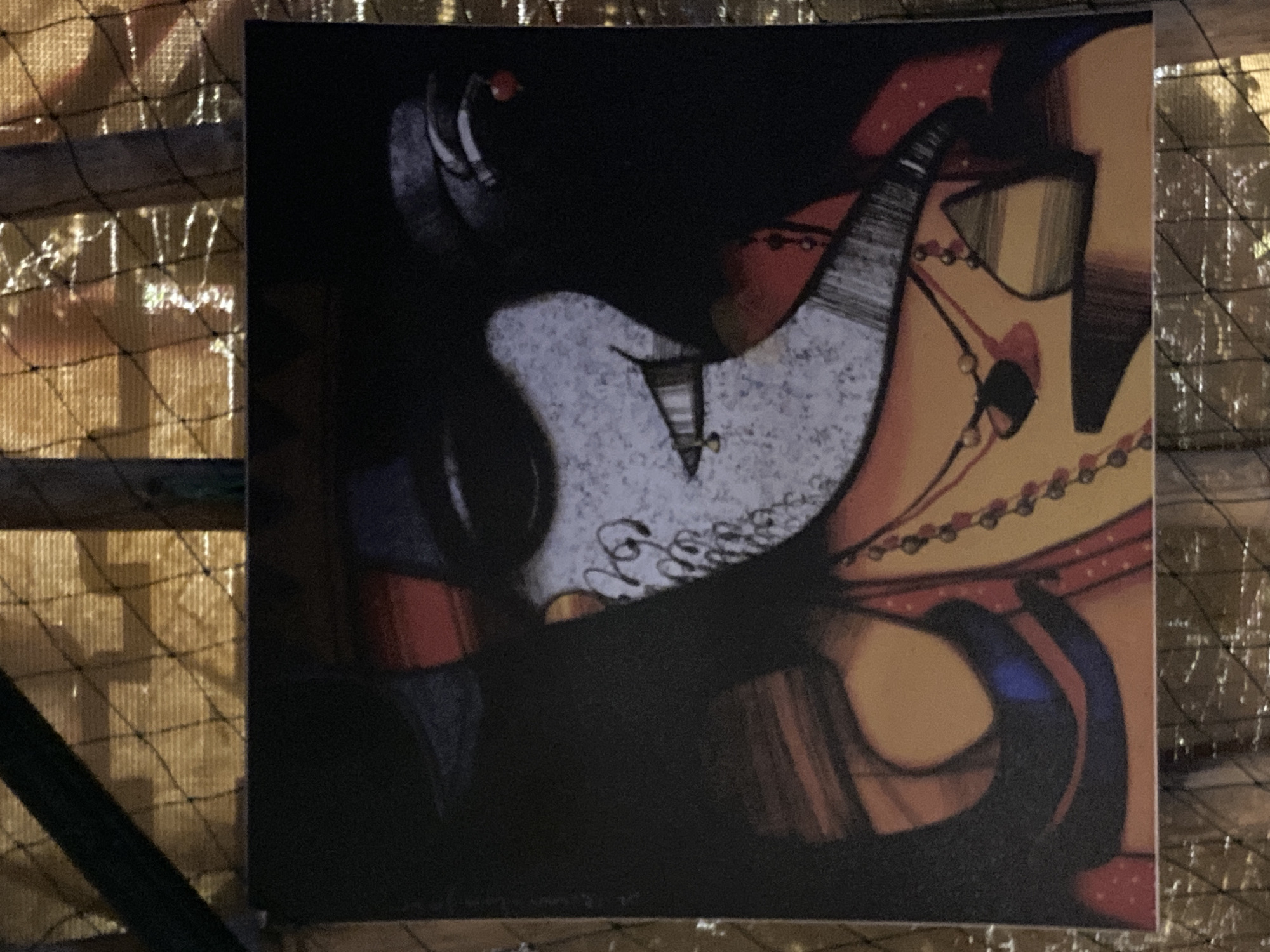 The image is a photograph of an abstract artwork hanging on a structure, possibly a fence, made of wooden planks with a black net. The artwork features stylized figures or faces with dark colors dominating the composition. One figure is painted with a light grey mottled texture, while another has a yellowish complexion and red accents. The artwork includes dark lines and shapes. The photograph was taken in Phu Thuy Ward, Vietnam.