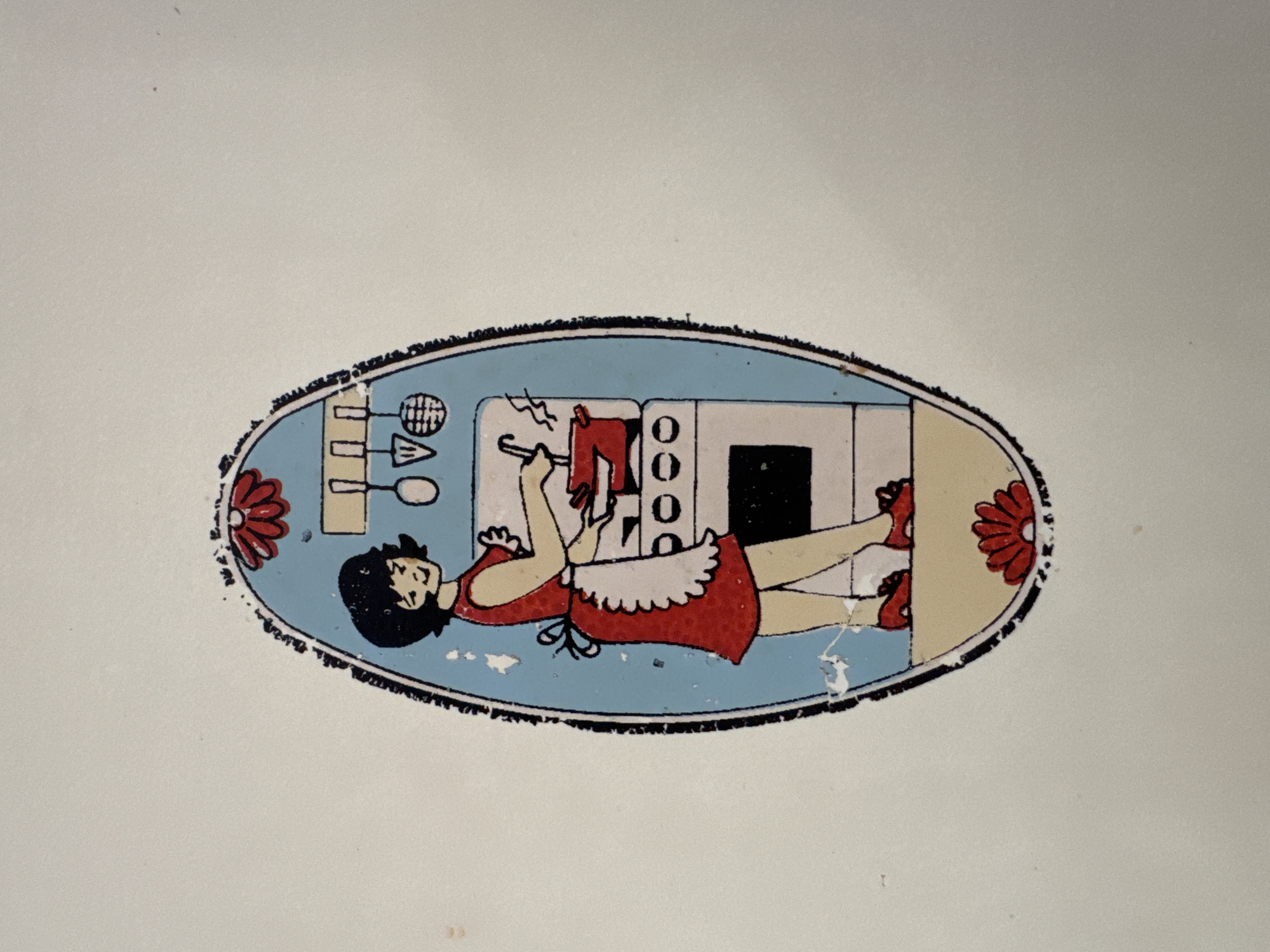 This is a close-up, rotated image of a decorative oval sticker on a light-colored surface. The sticker features a cartoon illustration of a person, likely a woman, lying down in front of what appears to be a kitchen stove. She has black hair and is wearing a red dress with a white ruffled apron. Her arms are raised, and she is holding a red object, possibly a kitchen utensil, near a stove with four circular burners. The stove has a white top and a black oven door. To the left of the stove are some kitchen-related icons, including what looks like a whisk or a rack, and some geometric shapes. The scene is set against a light blue background within the oval. Two red floral designs, one on the left and one on the right edge of the oval, add a decorative element. The illustration style is simple and somewhat retro. There is visible wear and tear on the sticker, with chipping and peeling paint along the edges. The overall impression is of a charming, domestic scene. No visible text is present in the image. The image is oriented such that the bottom of the oval sticker is to the right.