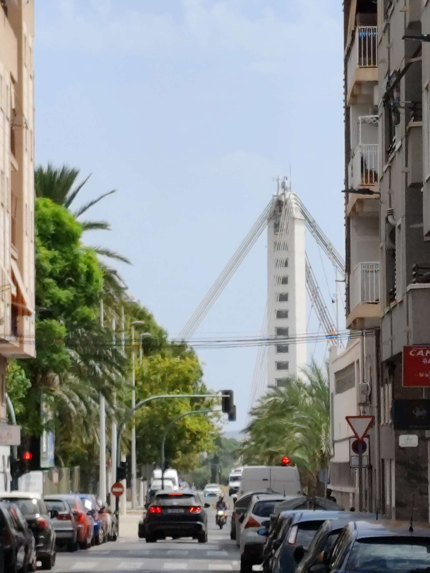 The image depicts a street scene in an urban environment. On the right side of the frame, a multi-story building with balconies and visible utility lines occupies a significant portion of the view. A red sign with white lettering is affixed to the building's facade. On the left side, another building is partially visible, with windows and balconies. Between the buildings, a street is lined with parked cars, and several vehicles are in motion. A traffic light with a red signal is visible on the left. Palm trees and other greenery line the street, casting shadows. In the background, a distinctive white suspension bridge with tall towers and cables is prominent against a pale blue sky. The scene appears to be a daytime view of a city street with a bridge in the distance.