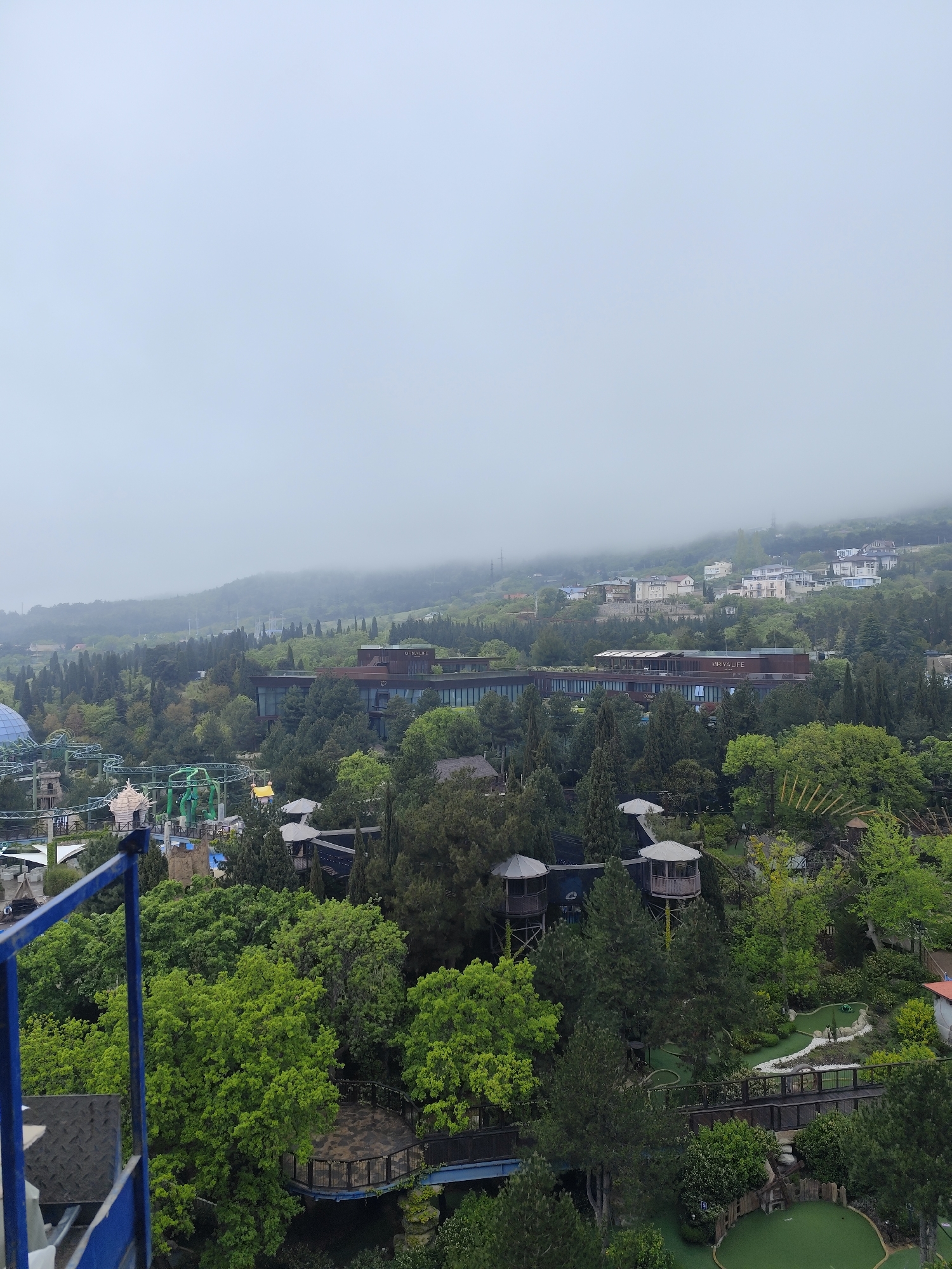 The image shows an aerial view of a resort area in Симеизский поселковый совет, Ukraine, under foggy conditions. The foreground features dense greenery, including trees and shrubs. There are small gazebo-like structures elevated on stilts amongst the trees. To the right, there's a mini-golf course with green synthetic turf. In the background, a large, multi-story building with a modern design stands out against the hazy backdrop of a mountain range partially obscured by fog. A rollercoaster is visible on the left side of the image. The sky is overcast and gray, suggesting a cloudy day. A blue metal structure is visible in the lower left corner of the image, likely a part of the observation platform.