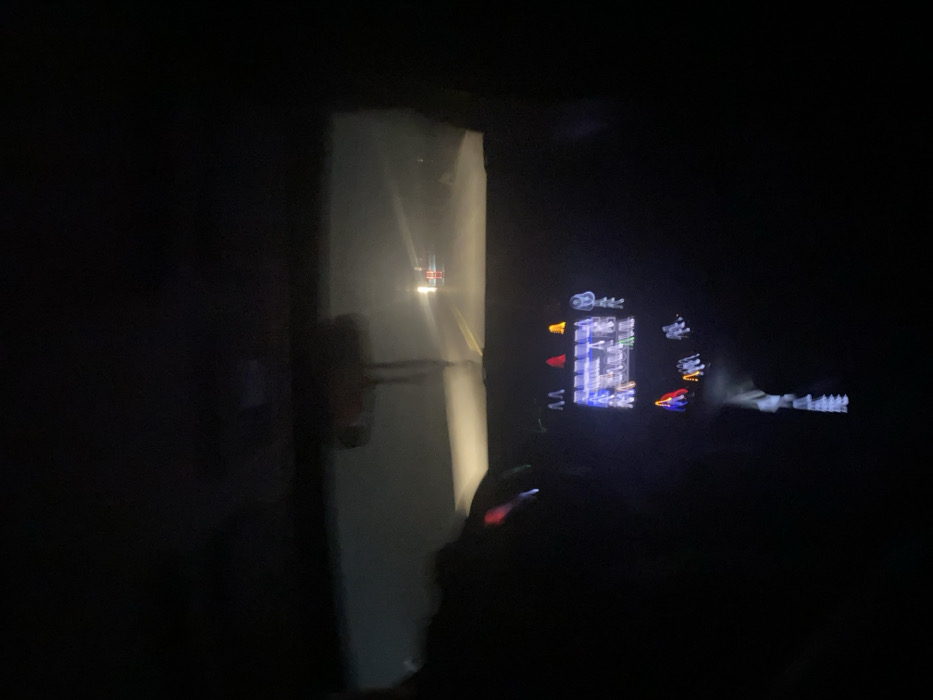 The image depicts a nighttime scene from inside a vehicle traveling on a road in Sharq tongi, Uzbekistan. The perspective is from the driver's or passenger's seat, looking forward through the windshield. The environment outside is dark, with limited visibility. The vehicle's headlights illuminate a portion of the road ahead, revealing faint white lane markings. In the distance, two bright, diffused light sources are visible, accompanied by vertical red reflective elements, suggesting another vehicle or construction hazard.

Inside the vehicle, a portion of a human arm is visible on the left, gripping a steering wheel, indicating the presence of a driver. The dashboard's center console is illuminated, displaying a screen with various glowing icons and symbols. These include red and orange/yellow triangular warning lights and other blue/white symbols, likely related to vehicle status or infotainment. A blurry object hangs from the rearview mirror. The overall scene suggests vehicular travel during dark conditions, possibly with some atmospheric haze affecting visibility.