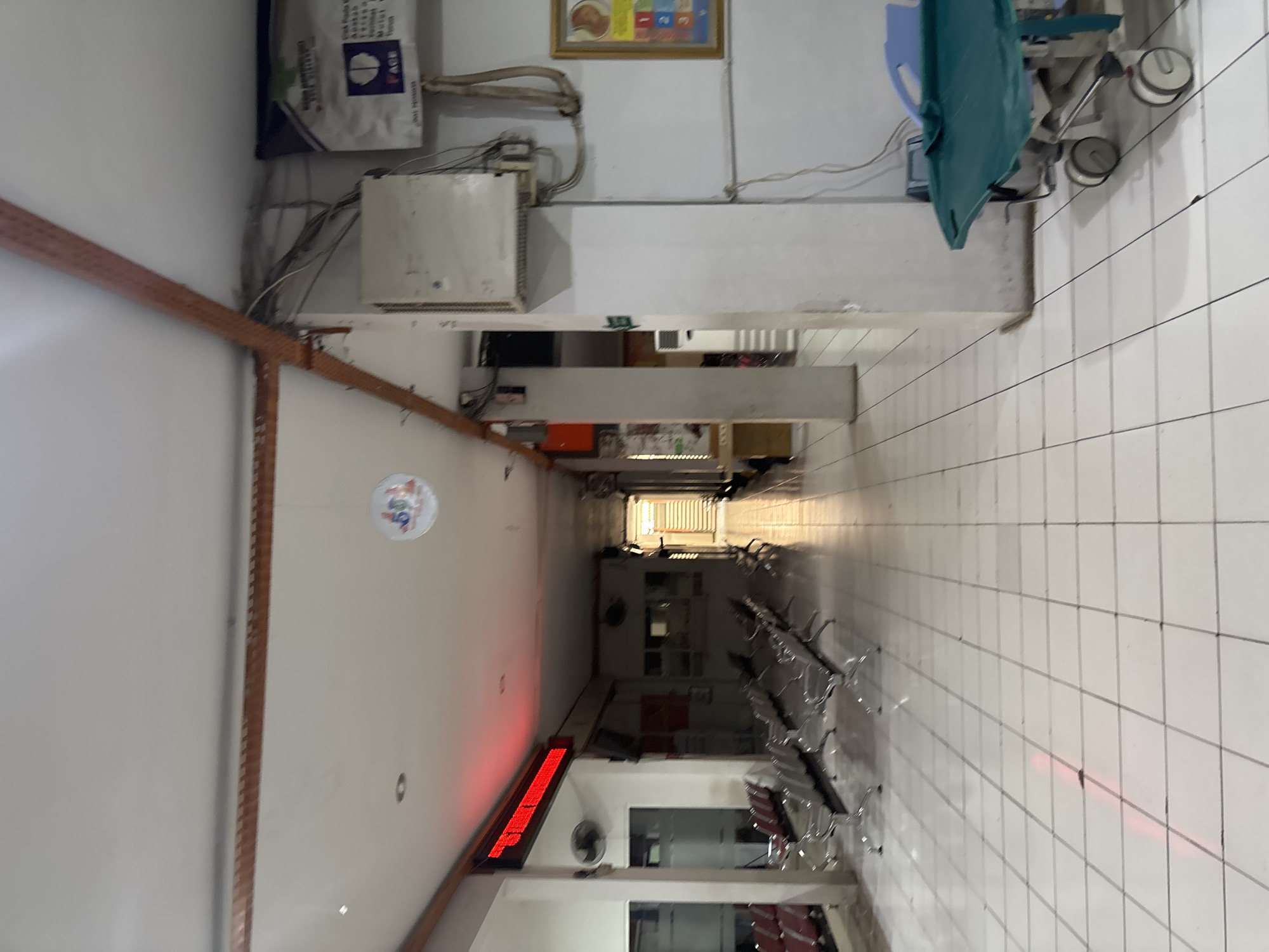 The image depicts a brightly lit, tiled hallway within what appears to be a hospital or clinic waiting area in Cirebon, Indonesia. Rows of empty metal waiting chairs line the left side of the hallway, facing a large red digital display board. A fan is mounted on the wall near the display. The hallway extends into the distance, with more seating visible. On the right side of the hallway, a medical stretcher with a green padded surface and wheels is parked near a wall. Above the stretcher, an electrical junction box is mounted on the wall, with various wires connected. A framed picture, possibly a chart with numbers and illustrations, hangs on the wall further down the hallway. The ceiling features exposed electrical conduits and a circular hanging decoration. The floor is covered in white tiles with dark grout lines. The overall impression is one of a clean, functional, and currently unoccupied public indoor space.