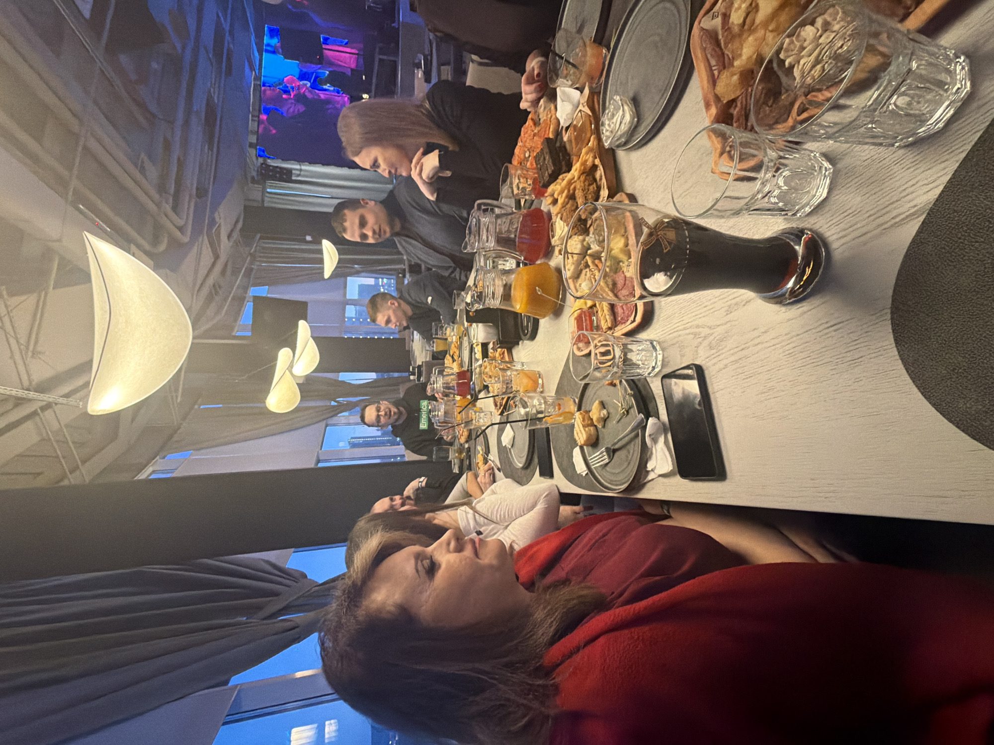 This image captures a social gathering of several people seated around a long table in what appears to be a modern restaurant or bar in Krasnoyarsk, Russia. The perspective is from the side of the table, looking along its length.
In the immediate foreground, a woman with long, dark hair, wearing a red top, is seen in profile, looking to her right with a neutral expression. Behind her, another woman in a white top is partially visible. Further down the table, a man with a dark beard is visible, and beyond him, a smiling man wearing a dark top with the word "Emerica" printed on it looks towards the camera. On the opposite side of the table, a woman with long brown hair, dressed in a dark top, is leaning on the table, while a man in a dark shirt sits across from her. More individuals are seated further down the table, engaged in conversation or enjoying the meal.
The table is laden with a variety of food and drinks. Numerous platters hold an assortment of appetizers including what looks like charcuterie (salami, cheese), various types of bread, crackers, nuts, and other small bites. Glasses filled with different beverages, such as beer in a tall glass, juices (orange and red), and other cocktails, are present. Plates, cutlery, and mobile phones are also scattered on the light-colored wooden tabletop.
The setting features modern interior design with unique, boat-shaped pendant light fixtures hanging from the ceiling. Large windows reveal an urban cityscape at dusk or night, with city lights visible against a dark blue sky. In the background, a large screen displays abstract, colorful visuals, possibly from a performance or video art. The atmosphere appears relaxed and social. The visible text is "Emerica" on a man's shirt.