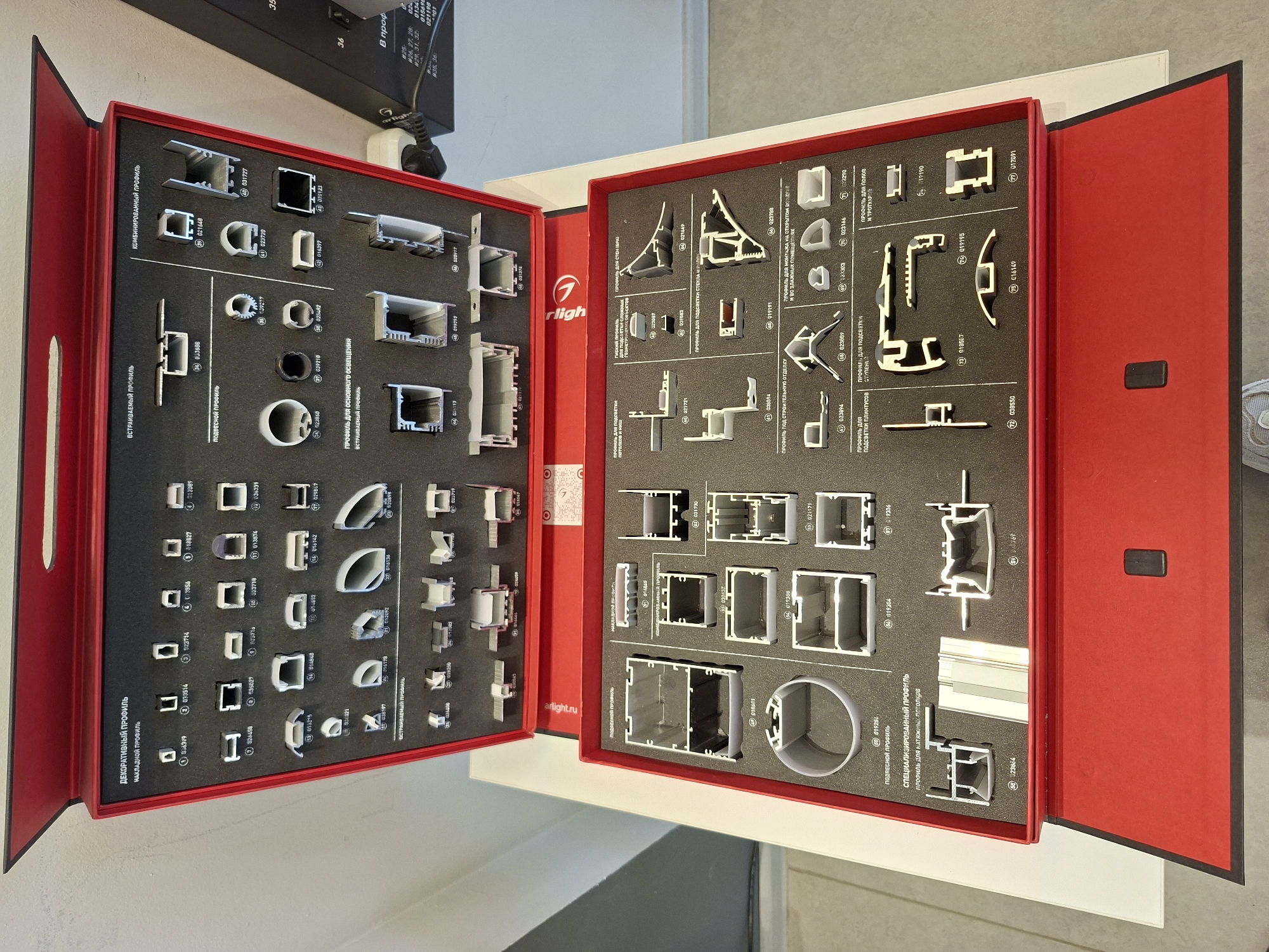 This is a media file of a display case filled with aluminum profiles. The display case is red with black interior foam inserts, each cut to hold different shapes of aluminum extrusions. The profiles are arranged in sections with Russian text labels indicating their purpose, such as "DECORATIVE PROFILE," "BUILT-IN PROFILE," "COMBINED PROFILE," "SUSPENDED PROFILE," and "SPECIALIZED PROFILE." Each profile is numbered and some have alphanumeric codes printed on them. The display case appears to be open, revealing two hinged sections filled with various profile samples. A logo that reads "rligh" and "artight.ru" is visible on the display. The background suggests an indoor setting, possibly a showroom or trade fair booth, with a glimpse of a black electronic device with Cyrillic text and numerical labels in the upper left corner, and a grey floor and wall in the background. The lighting is bright and even, suggesting an indoor commercial or industrial environment. There are no people visible in the image. The overall impression is of a product catalog or sample display for architectural or construction materials.