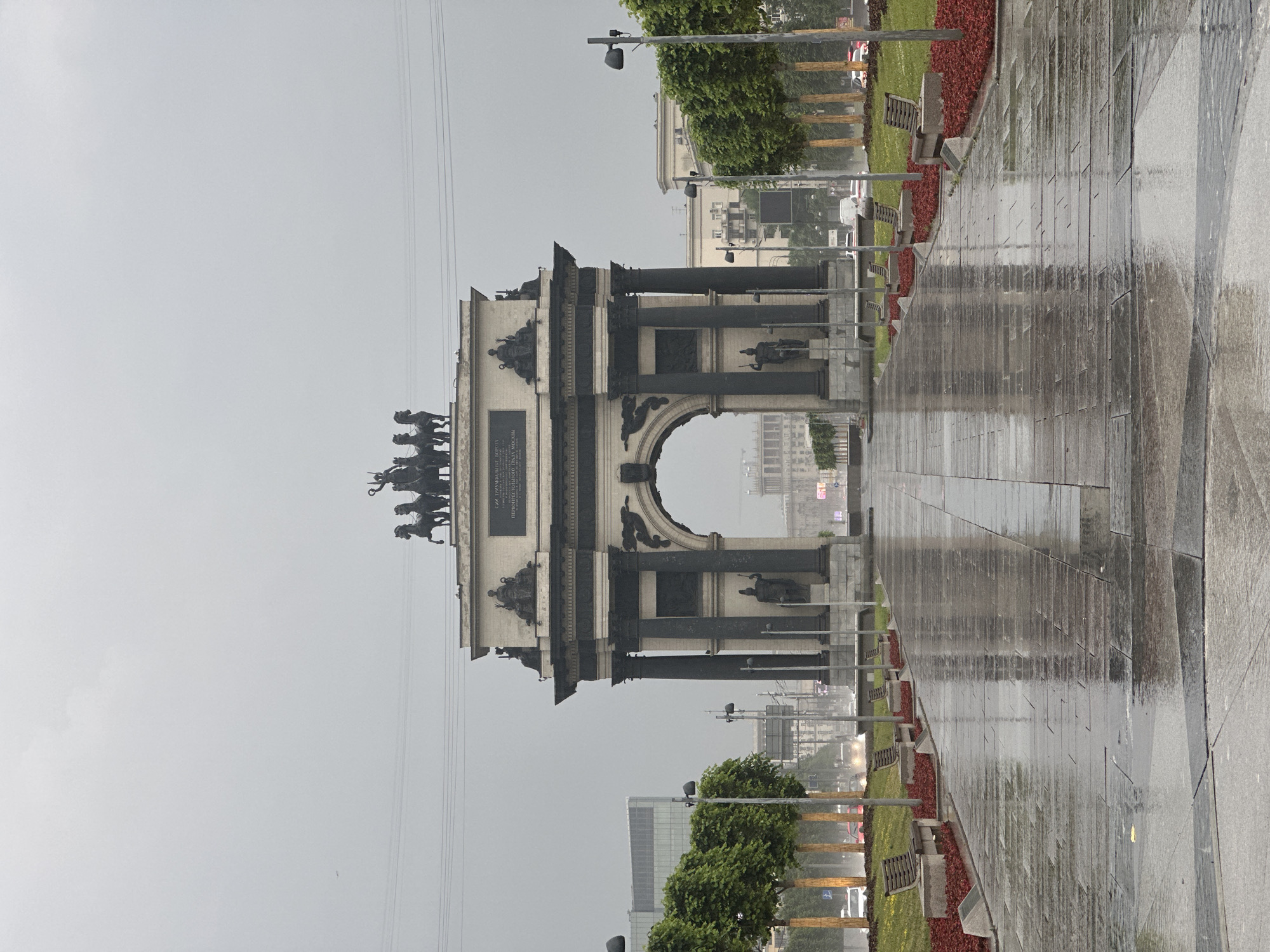 The photograph depicts the Triumphal Arch in Moscow, Russia, on an overcast, rainy day. The arch, a prominent architectural landmark, stands in the center of the frame, its imposing structure dominating the scene. The sky is a uniform gray, with faint power lines stretching across the upper portion of the image. The ground in the foreground is a wet, reflective pavement, suggesting recent rainfall. Rows of green trees line the pathway leading to the arch, their foliage appearing damp. Several park benches are visible along the sides of the path, interspersed with beds of red mulch and green grass. In the distance, through the archway, more buildings and city elements are visible, though blurred by the atmospheric conditions. The overall mood of the image is somber and tranquil, emphasizing the architectural grandeur of the arch amidst a quiet, rainy urban setting.