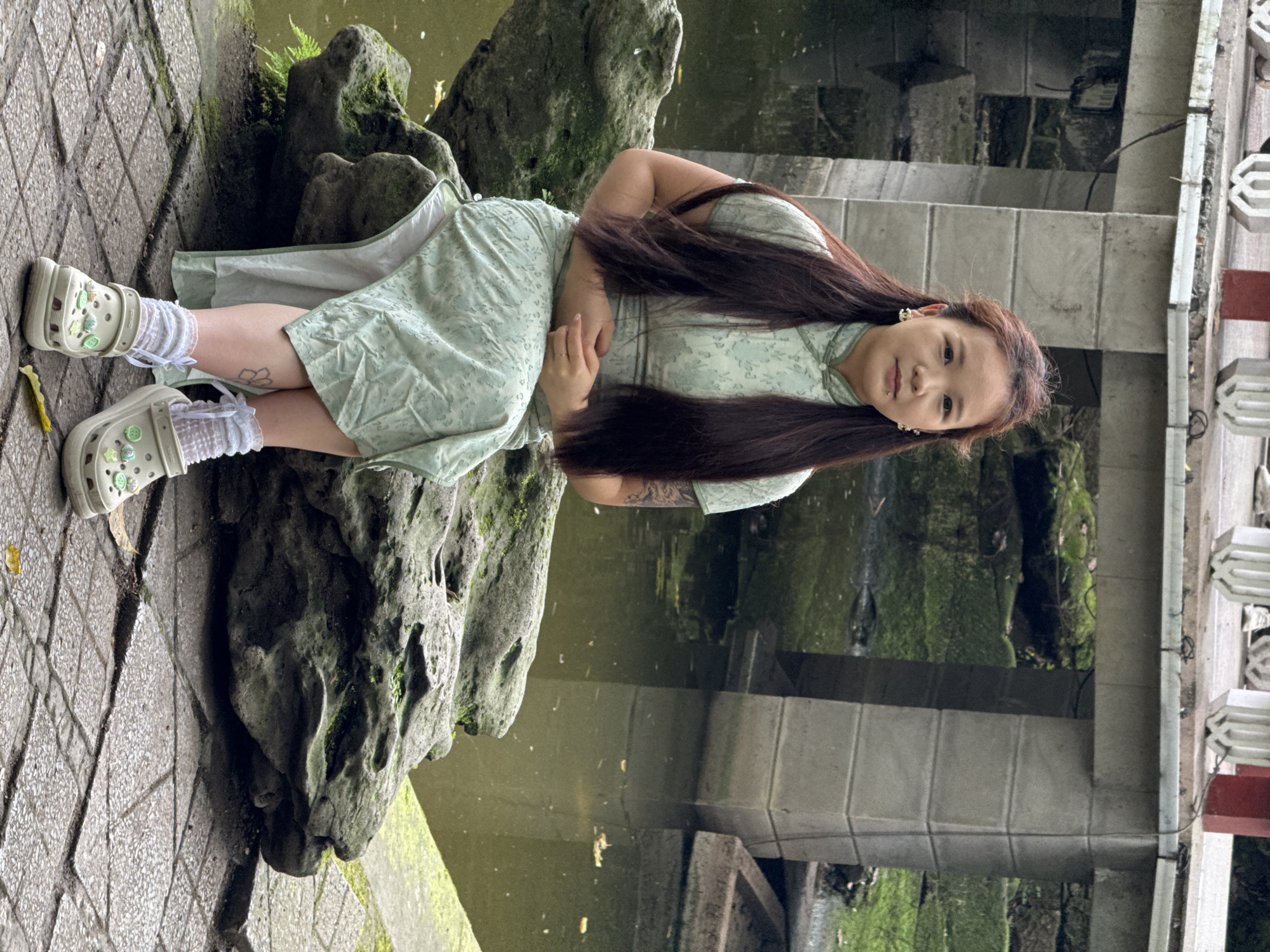 A woman with long dark hair is seated sideways on a rock near a pond in Yuzhong District, China. She is wearing a light green dress and light tan Crocs with white socks. She is posed facing the camera. The background includes a pond, bridge, mossy rocks, and a stone-paved area.