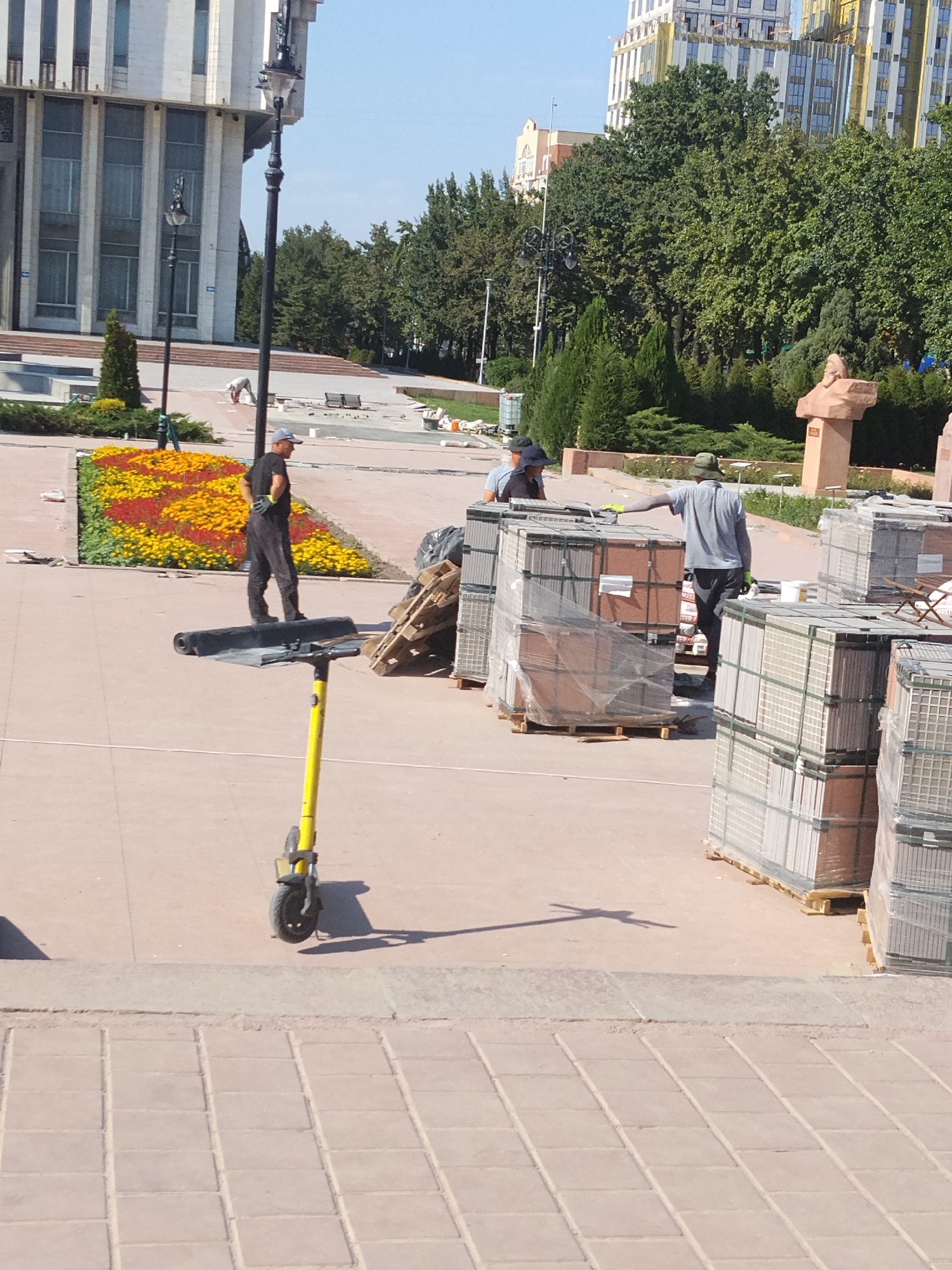 The image depicts a public area in Bishkek, Kyrgyzstan, possibly a park or a plaza, undergoing renovation or construction. In the foreground, a yellow electric scooter is parked with its handlebars facing left. A person wearing black overalls and a cap stands on the scooter's platform, appearing to be taking a break. Behind them, a colorful flower bed with red and yellow flowers is visible.
Further back, several workers are engaged in tasks related to the construction. One individual in a grey shirt and green hat is seen near stacks of materials, possibly tiles or building blocks, wrapped in plastic and arranged on wooden pallets. Another person in dark clothing and a hat is partially visible among the materials. In the background, a large, modern building with a light-colored facade and numerous windows dominates the skyline, along with lush green trees and some construction scaffolding on another building. The ground surface is paved with light-colored tiles. The sky is clear and blue, suggesting a sunny day.