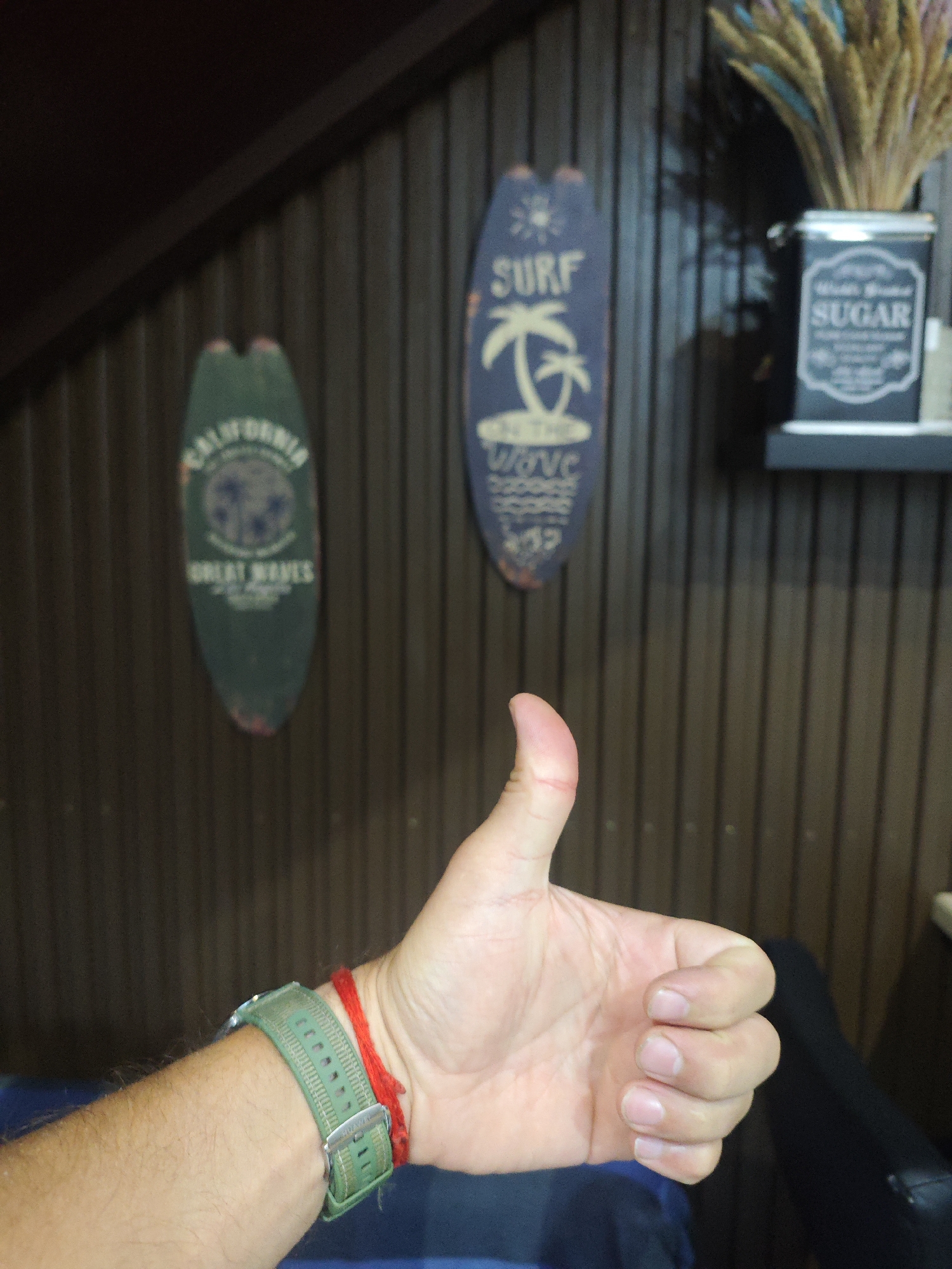 The image shows a person's hand giving a thumbs up gesture. The hand has a green watch with a red string tied around the wrist. In the background, there are two decorative surfboards mounted on a wooden wall. One surfboard is blue and features a palm tree and the word "SURF." The other surfboard is green and has text that appears to say "CALIFORNIA" and "GREAT WAVES." To the right of the surfboards, a dark tin with the word "SUGAR" is visible on a shelf, accompanied by a bouquet of dried plants. The scene appears to be indoors. The location of Chaikovsky, Russia is not discernible from the image.