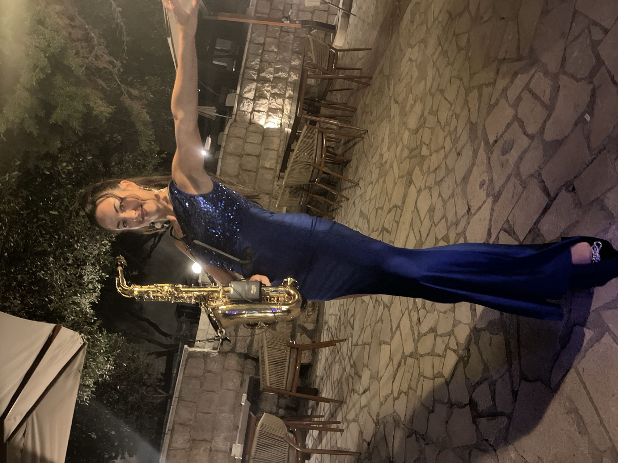A woman in a navy blue, sequined, sleeveless, floor-length dress and black high-heeled shoes stands in an outdoor courtyard at night, holding a golden saxophone with a black microphone attached. She is facing the camera, smiling, with her left arm extended to her right, her right arm bent and holding the saxophone. The courtyard has a flagstone floor and is surrounded by a stone wall. In the background, there are tables and chairs, an umbrella, and trees. The scene appears to be in the city of Pečurice, Montenegro.