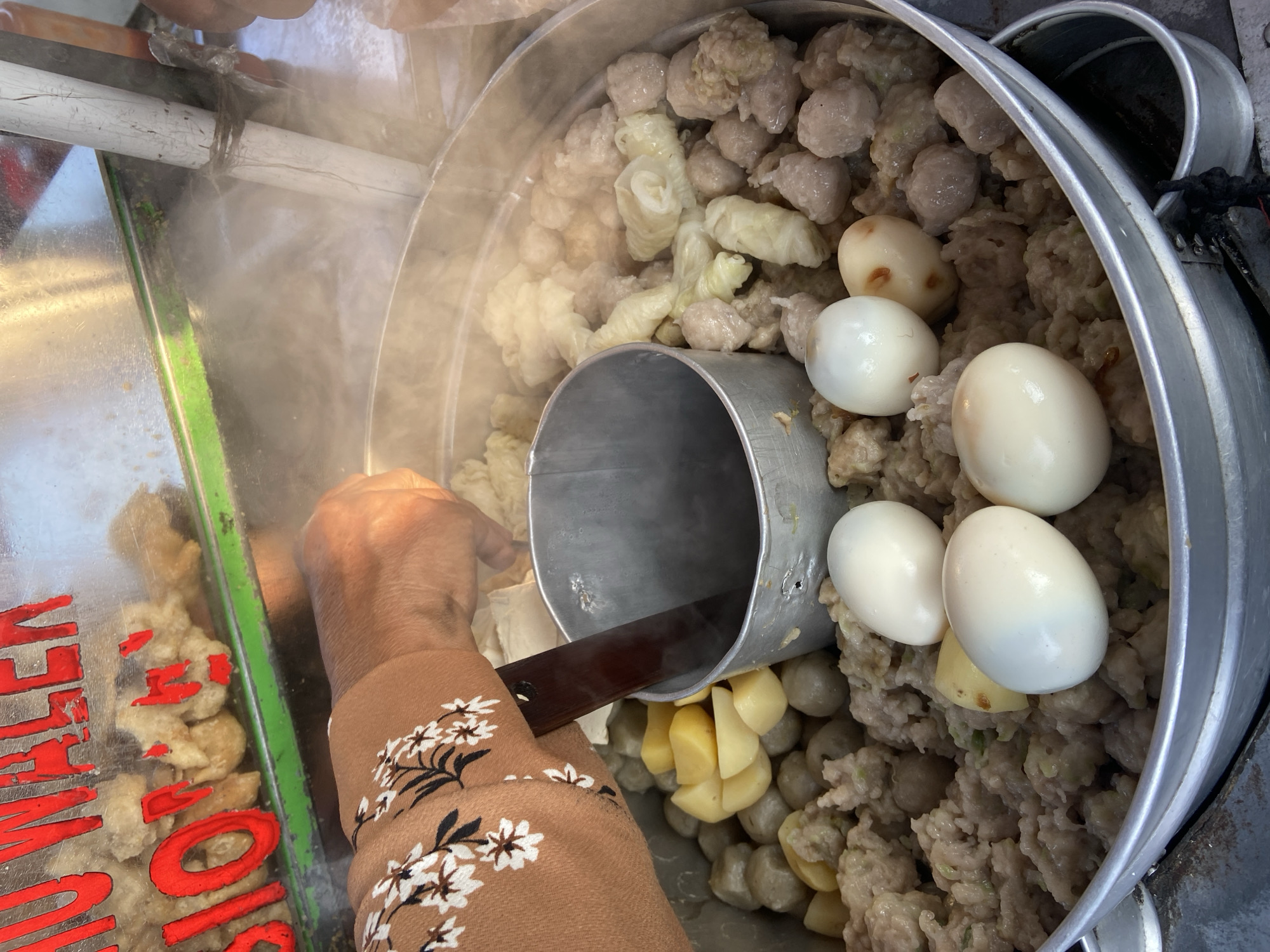 An overhead shot captures a street vendor in Tanjungrejo, Indonesia, preparing a dish from a steaming metal pot. The vendor, wearing a brown blouse with a floral pattern, is using a dark wooden utensil to stir the contents. The pot is filled with a variety of food items including meatballs, boiled eggs, and chopped potatoes, all covered in steam. In the background, a glass display case shows more food items and red lettering that is partially obscured. The scene appears to be outdoors, likely a busy street given the context of a street food vendor.