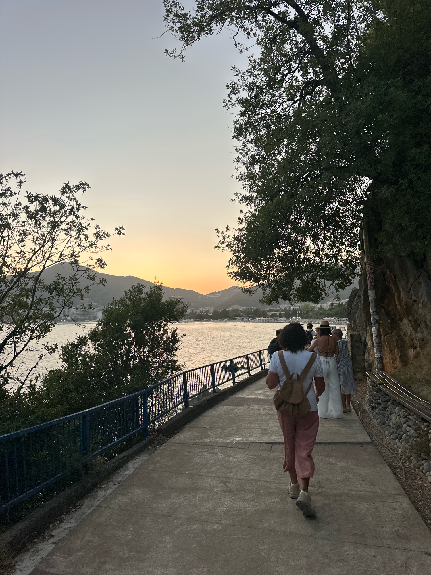 An exterior, eye-level, medium shot captures a group of people walking along a paved pathway beside a body of water, likely the Adriatic Sea, as the sun sets over a distant mountainous coastline near Budva, Montenegro. The pathway is bordered on the left by a blue metal railing and lush green foliage, and on the right by a rocky cliff face partially covered in vegetation. A prominent tree with dark green leaves extends from the upper right corner of the frame, casting shadows onto the scene. In the foreground, a woman from behind wears a white t-shirt, a light brown backpack, and pink trousers. She is followed by other individuals, including a woman in a straw hat and a black top with white trousers, and another in a striped dress. The water reflects the soft, warm hues of the sunset, which paints the sky in shades of orange and yellow. Buildings and the beachfront are visible in the distance on the far side of the water. The overall atmosphere is serene, suggesting a leisurely evening stroll.