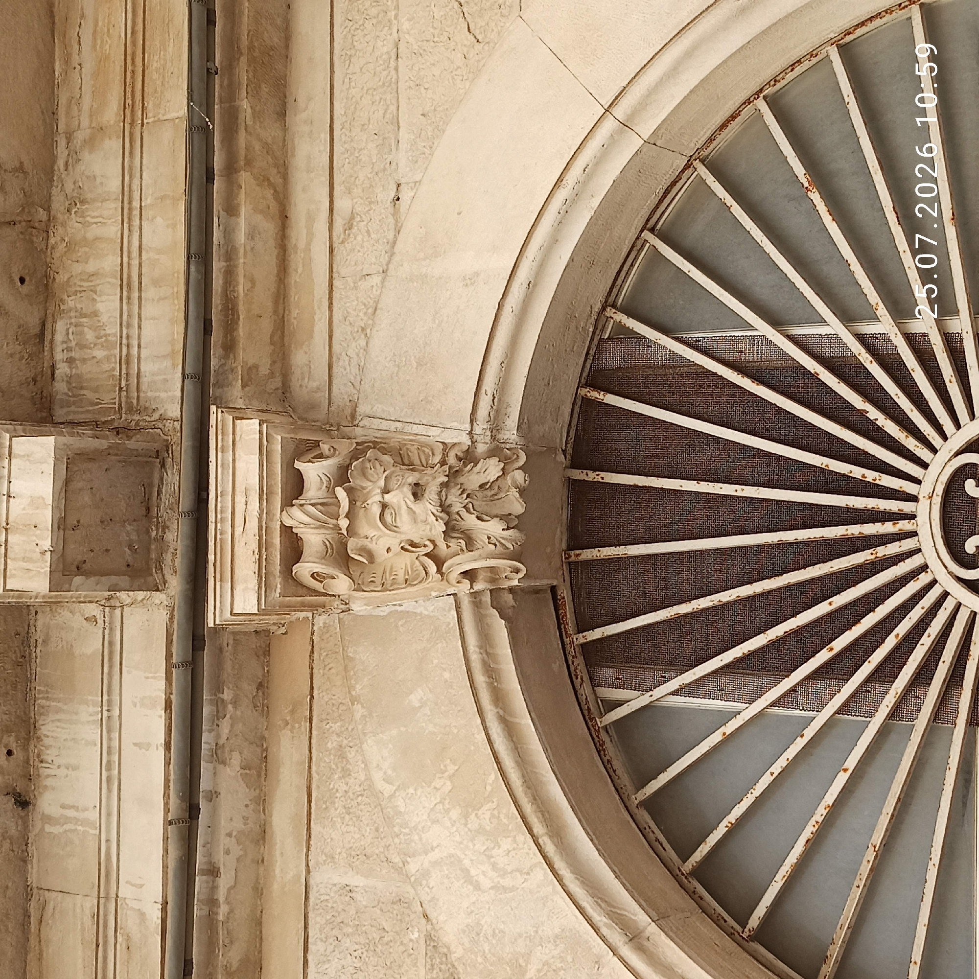 The image shows a close-up view of an architectural detail above a semicircular window in Ruvo di Puglia, Italy. The window has a white, radial metal grate and is partially covered by a dark mesh screen. Above the window, a stone corbel is carved into the face of a weathered stone building. The corbel features a grotesque face with leafy hair and a beard, in a style reminiscent of Baroque or Renaissance ornamentation. The building's facade is constructed of large, roughly hewn stone blocks, with visible signs of age, including water stains and chipped plaster. A dark pipe runs horizontally across the upper part of the facade, secured by metal brackets. The overall impression is of historical architecture in a state of preservation, with the ornate carving adding a distinctive character to the building. The date stamp "25.07.2026 10:59" is visible in the bottom right corner of the image, indicating when the photograph was taken.