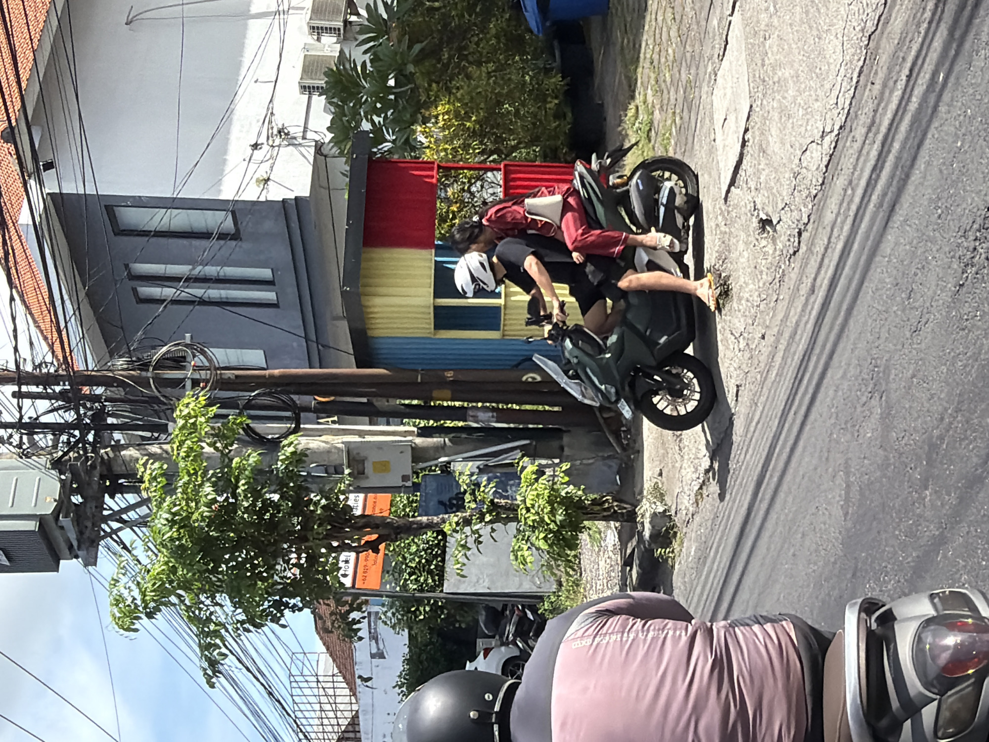 The image shows a street scene in North Kuta, Indonesia. Two people are on a scooter. The person driving the scooter wears a black t-shirt, black shorts, and a white helmet. A passenger, dressed in red pajamas, is seated behind the driver. Another scooter is in the foreground, partly obstructing the view. The scene includes buildings, trees, and overhead power lines. A colorful, corrugated metal fence, with sections of red, yellow, and blue, stands near the scooter. The road shows signs of wear and repair.