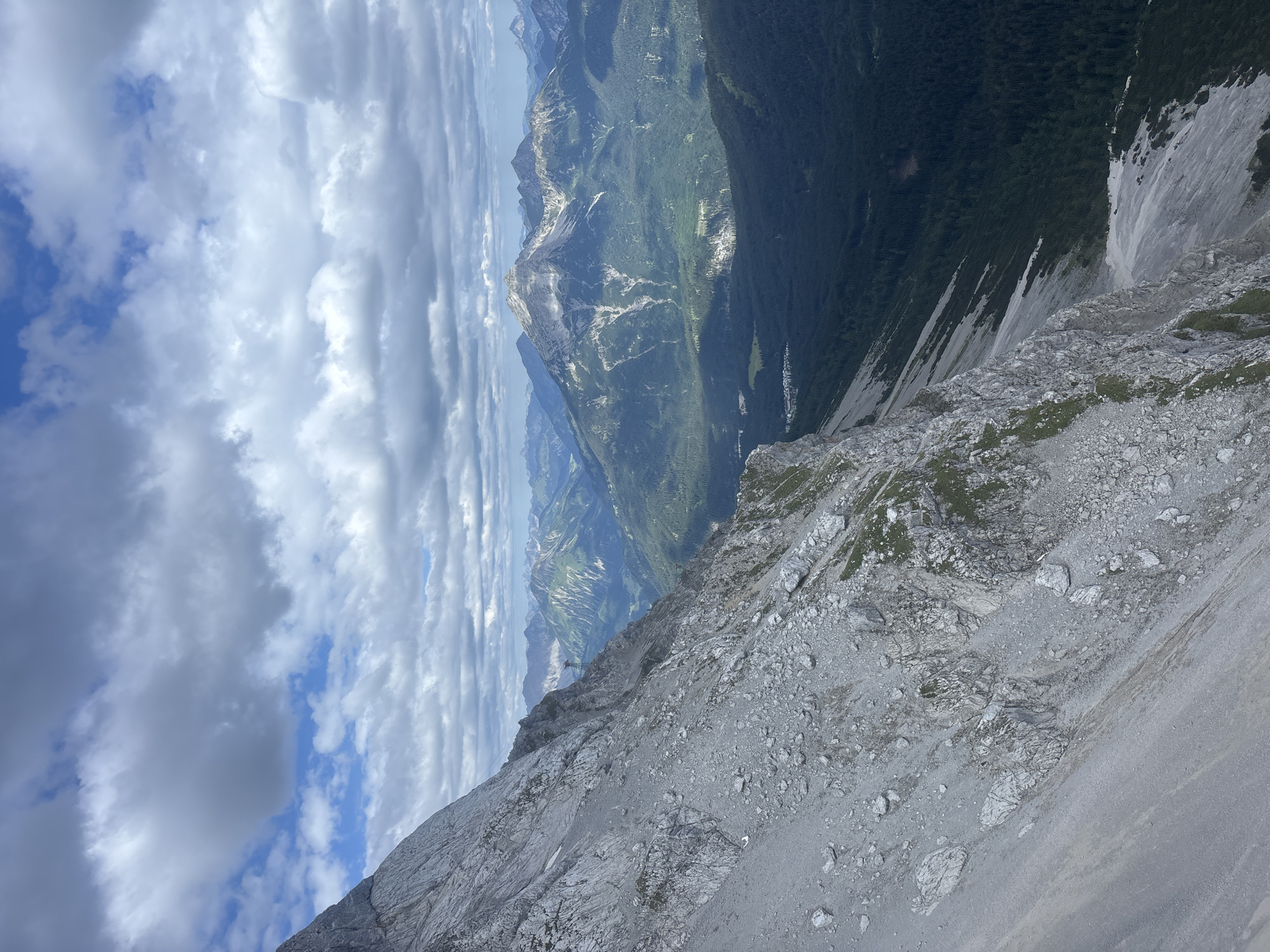 This image, taken from a high vantage point in the vicinity of Grainau, Germany, captures a vast mountainous landscape. In the foreground, a steep, rocky mountainside dominates the left side of the frame, its grey scree and boulders stretching downwards at a sharp angle. Patches of green vegetation are sparsely present on the rock face.
Beyond the immediate mountainside, the scene opens up to a sweeping view of several mountain ranges. The middle ground features a broad valley filled with lush green forests. A small cluster of buildings, indicative of a village, is visible in the valley floor. Further in the distance, progressively higher and more rugged mountain peaks rise, some displaying patches of snow or light-colored rock. The furthest peaks fade into a hazy blue, suggesting a considerable distance.
The sky above is a mix of dramatic clouds and patches of blue. Large, grey, cumulus clouds are present, with some appearing darker and more textured than others. Sunlight breaks through in various parts of the sky, casting shadows on the landscape below. The overall impression is one of immense natural scale and a sense of elevation. There are no people or discernible actions depicted in the image.