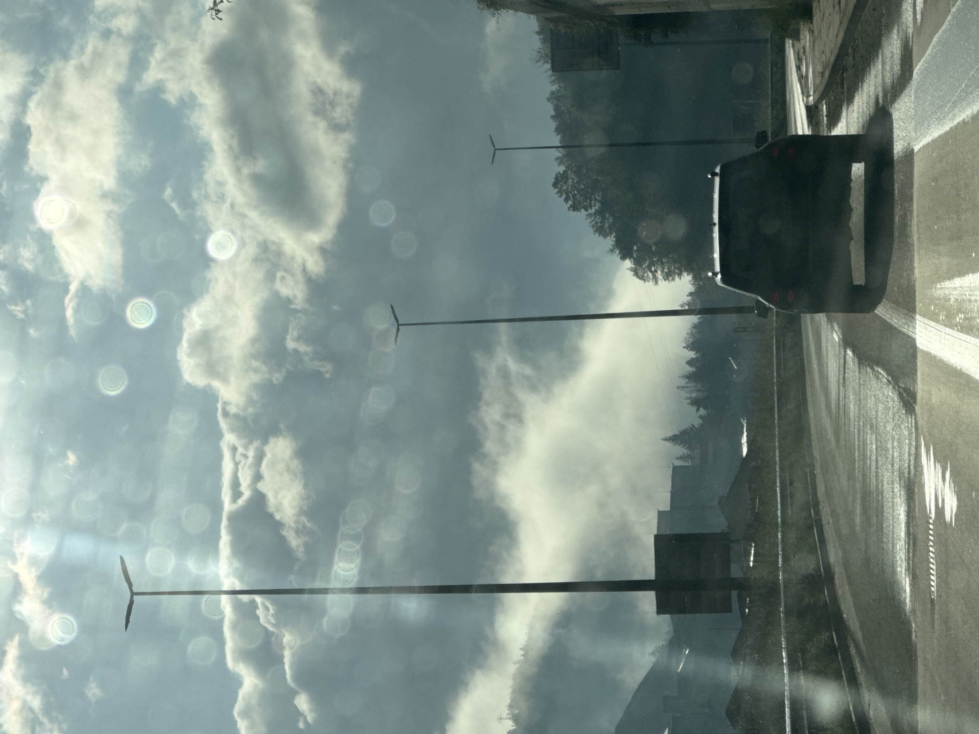 The image shows a view from inside a car traveling on a wet road in Smolyan, Bulgaria. The road is lined with street lights. A dark-colored vehicle is slightly ahead in the adjacent lane. Buildings and trees are visible on the side of the road. The sky is cloudy. Bokeh is visible on the windows.