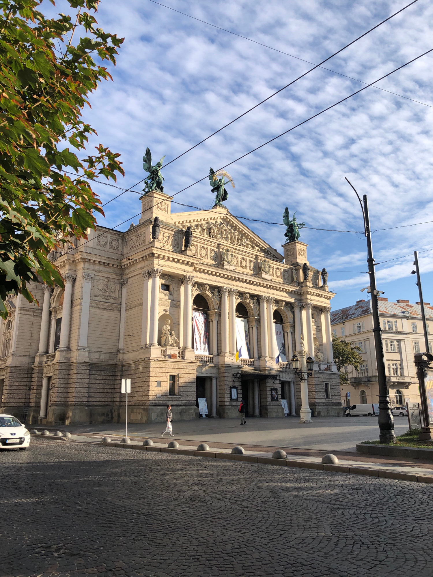 The image shows a wide shot of the Lviv Opera House, a grand neoclassical building in Lviv, Ukraine, situated in front of a partly cloudy blue sky. The building's facade is adorned with intricate sculptures and classical architectural elements, including columns and statues. A tree with green and brown leaves is visible in the upper left corner, partially obscuring the sky and the top of the building. In the foreground, a cobblestone street curves away from the viewer, with a white car parked on the left side. A pedestrian walks along the sidewalk in front of the opera house. The scene is illuminated by natural light, suggesting it was taken during the daytime. The overall atmosphere is serene and urban, capturing a moment of daily life in front of a prominent cultural landmark.