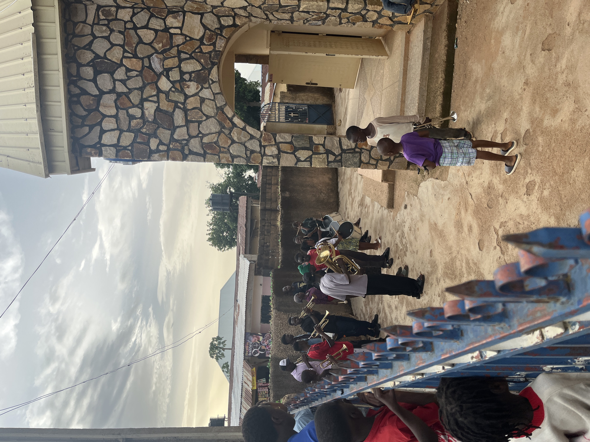 A medium shot depicts a brass band lined up along a dirt path, observed by children behind a painted fence, likely in Gura Top, Nigeria. The band consists of approximately ten individuals in various attire, some in white shirts, playing brass instruments. Two younger individuals stand on a raised platform near a stone building with an arched entrance. The setting is a sunlit day with partly cloudy skies.