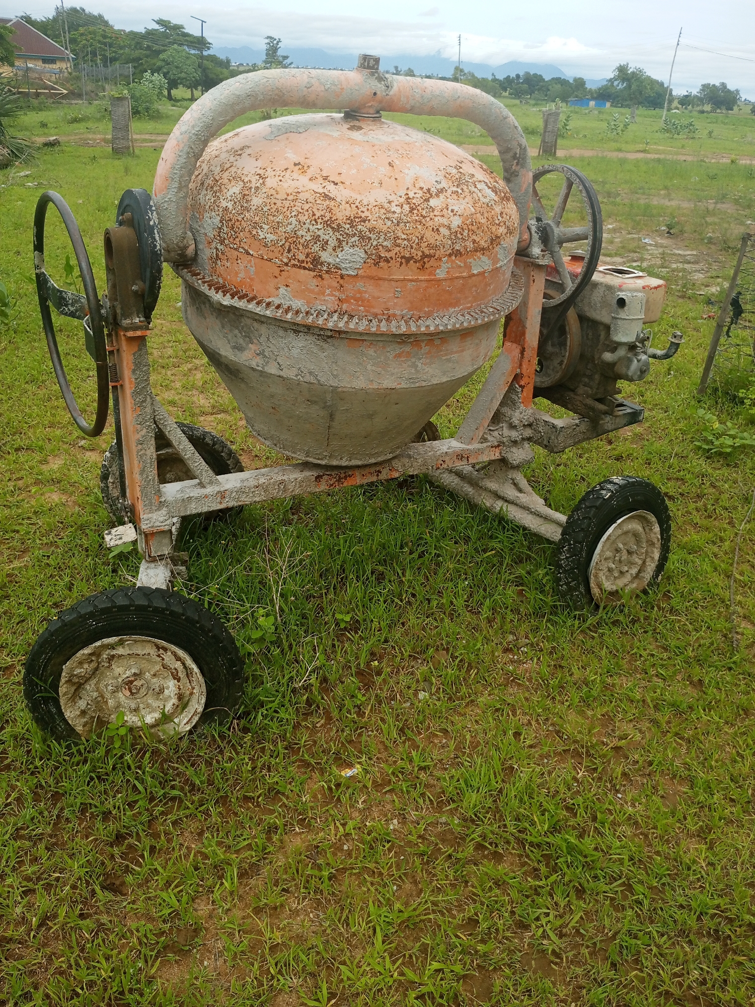 A rusted orange cement mixer sits on a grassy field in Jalingo, Nigeria. The mixer is covered in dried cement, indicating recent use. It has four wheels, two of which are on the ground and appear to be flat. The mixer has a large drum that is tilted forward, and a handle on the side used to tilt it. A small engine is attached to the back of the mixer. The background shows a few scattered trees and buildings, as well as a cloudy sky. The overall scene suggests a construction site in a rural or semi-rural area.