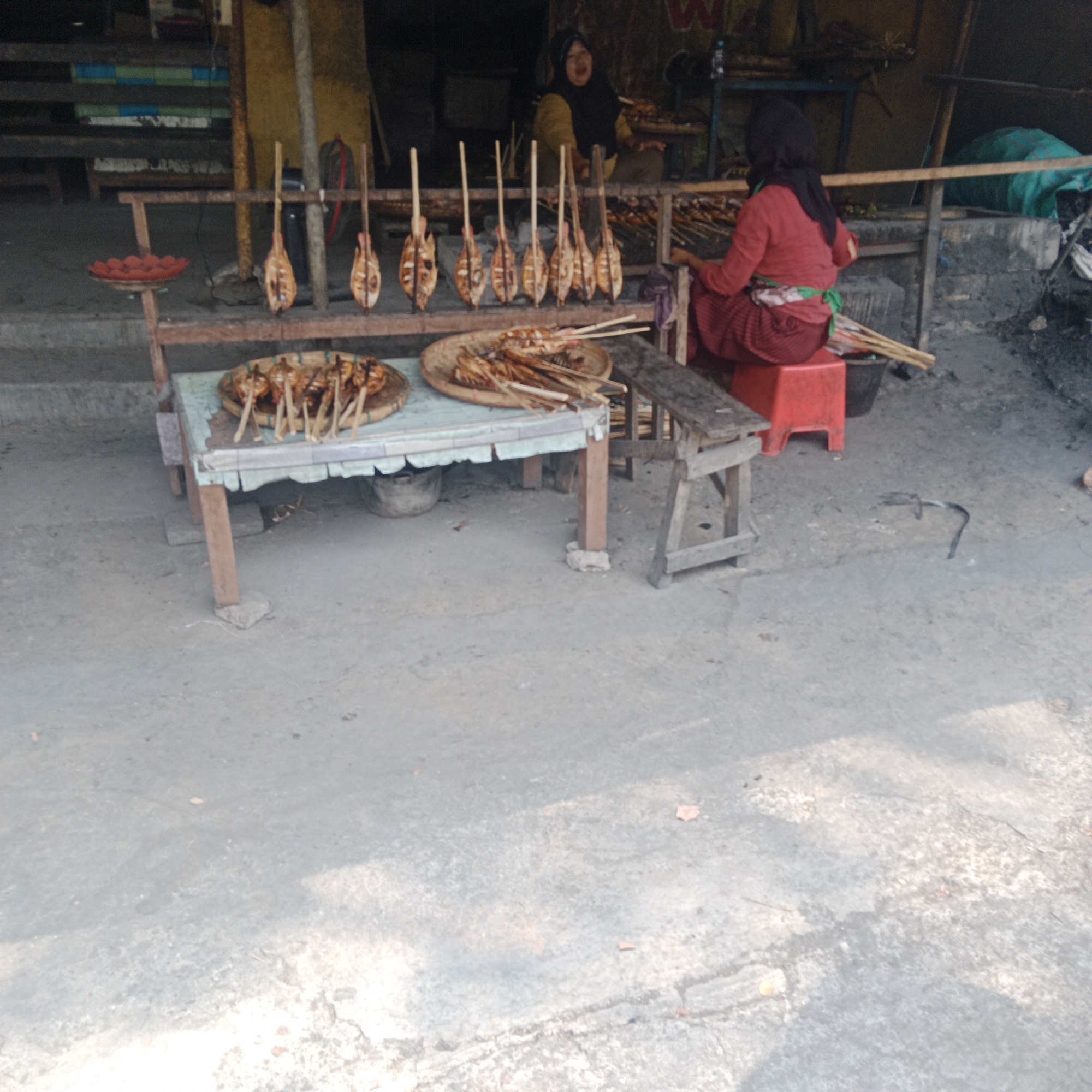 The image depicts an outdoor scene in Ngandul, Indonesia, where food is being sold from a makeshift stall. In the background, two women are present. One woman, wearing a dark headscarf and a yellow top, appears to be interacting with a customer or preparing food. The other woman, wearing a red long-sleeved top and a headscarf, is seated on a red plastic stool, also appearing to be involved in the food preparation or sale.

In the foreground and midground, several food items are displayed. On a table covered with a light blue and white patterned cloth, two woven circular trays hold grilled food on skewers. One tray contains what appear to be fish, and the other contains what could be similar items or other types of skewers. In front of the table, a wooden structure with multiple skewers of grilled food is displayed horizontally on a wooden rack, with each skewer featuring a single piece of food that has been cut and prepared for grilling. Another rack, set up higher, displays several more identical skewers of grilled food. A small red dish containing what appear to be small, round red fruits or candies is also visible.

The environment appears to be an outdoor market or street-side stall. The ground is paved with concrete. The stall itself is constructed with wooden posts and beams, with a partial roof or awning visible in the background. Various other objects, including a black bucket and some materials that could be firewood, are present around the stall. The overall impression is one of a local food vendor preparing and selling grilled items in a casual setting.