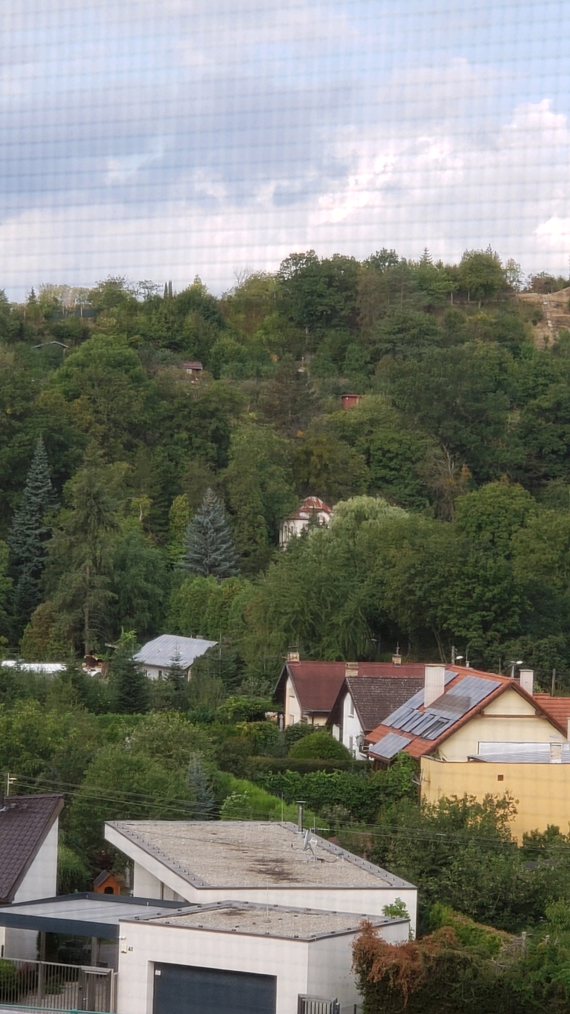 The image shows a view of a hilly, tree-covered landscape with several residential buildings visible amongst the foliage. The sky is overcast with gray clouds. In the foreground, there are several modern houses with flat roofs. One house on the right has a yellow facade and solar panels on its red-tiled roof. To the left, there is a white house with a garage and a smaller white building with a car shelter. Further up the hill, nestled within the trees, are more houses, some with red roofs and some with lighter colored roofs. A building with a domed roof is visible higher up on the hill, partially obscured by trees. The overall impression is a suburban or semi-rural residential area on a hillside.