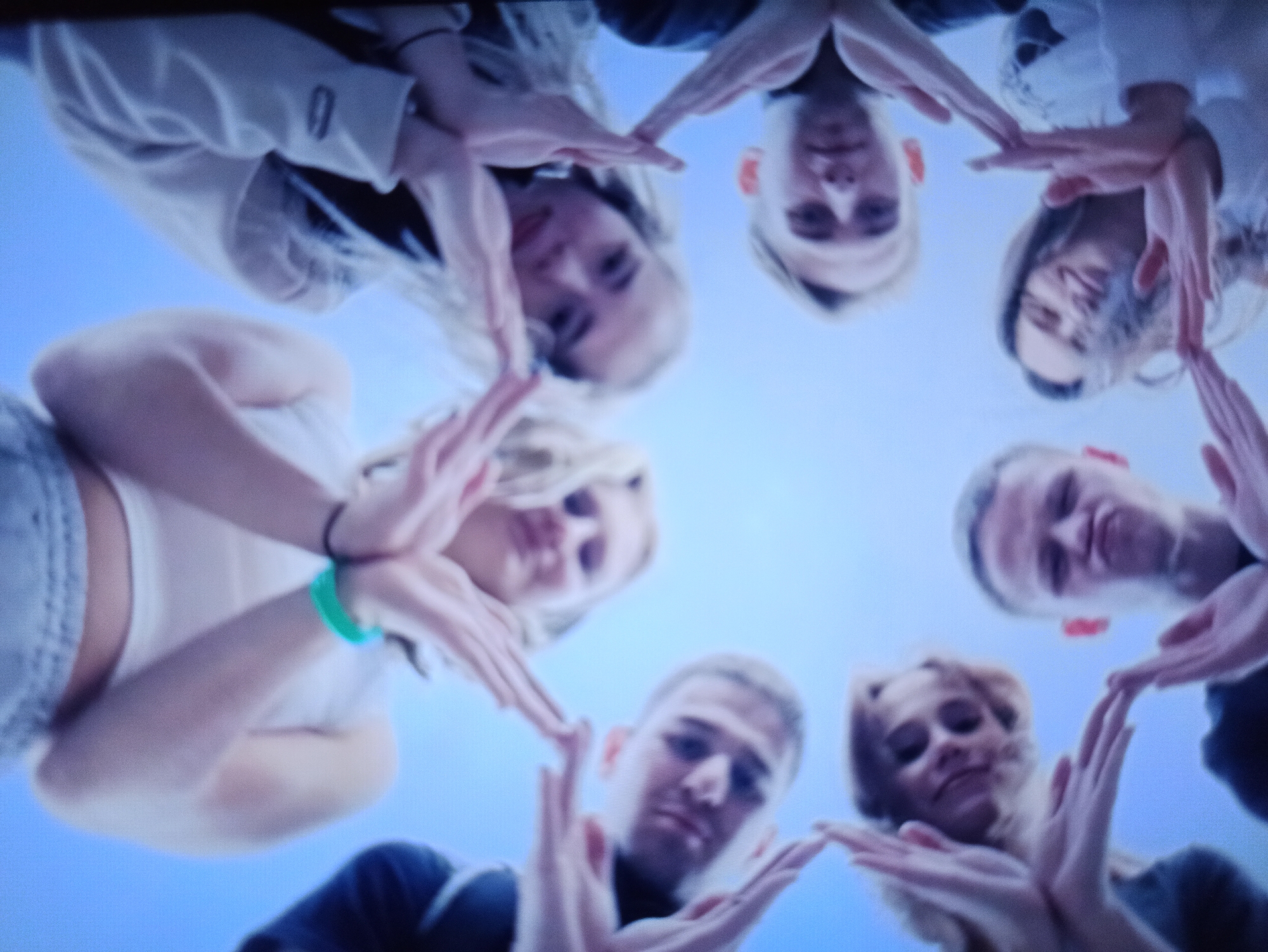 The image shows a low-angle, fisheye perspective of a group of eight young people forming a circle with their hands extended towards the center. They are all looking down at the camera, and their faces are partially obscured by their hands. The background is a clear blue sky. The people appear to be outdoors, but no specific city or location is identifiable. Their clothing suggests a casual setting, possibly a park or a gathering. The lighting is bright, indicating daylight.