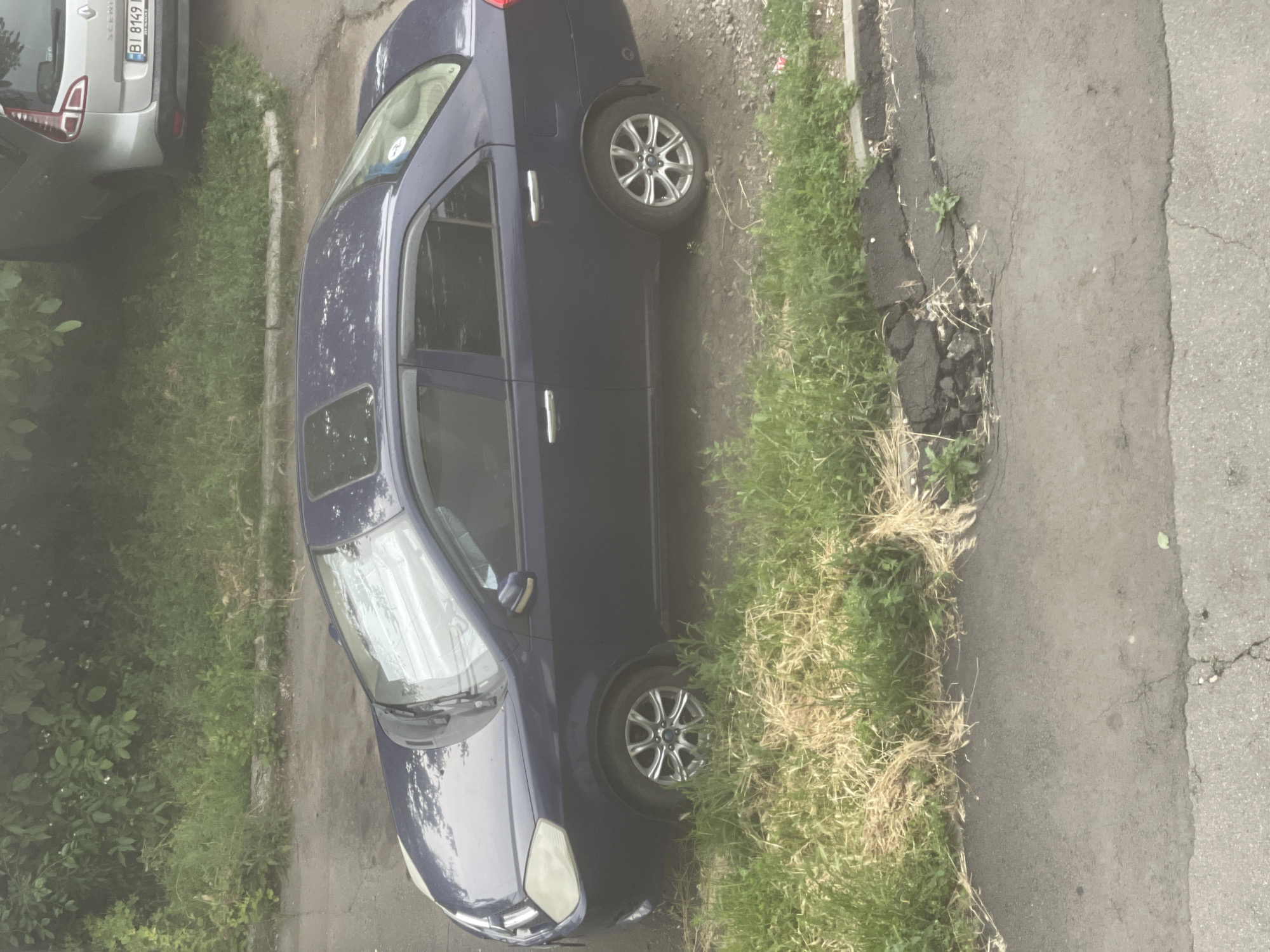 This is a portrait-oriented, somewhat hazy image of a dark blue sedan parked on the side of a road. The car is the primary object, taking up most of the frame. It appears to be a sunny day, judging by the reflections on the car and the bright green of the grass. The car is parked next to a strip of overgrown grass and weeds, which is adjacent to an asphalt road. The road shows signs of wear and tear, with cracks and a section that has broken away, exposing the ground beneath.
On the left side of the image, a portion of a silver car is visible, parked behind the blue sedan. The silver car has a license plate with white text on a blue background, reading "BI 6169 BI". There is also some text in a smaller font below that, appearing to be "RENAULT".
The blue sedan has a sunroof, and its windows are tinted. The car's wheels are visible, featuring silver alloy rims. The overall scene suggests an urban or suburban setting, possibly a residential street or a parking area. There are no people visible in the image, and the focus is on the parked vehicles and the environment. The image is slightly tilted, giving a dynamic perspective to the scene.