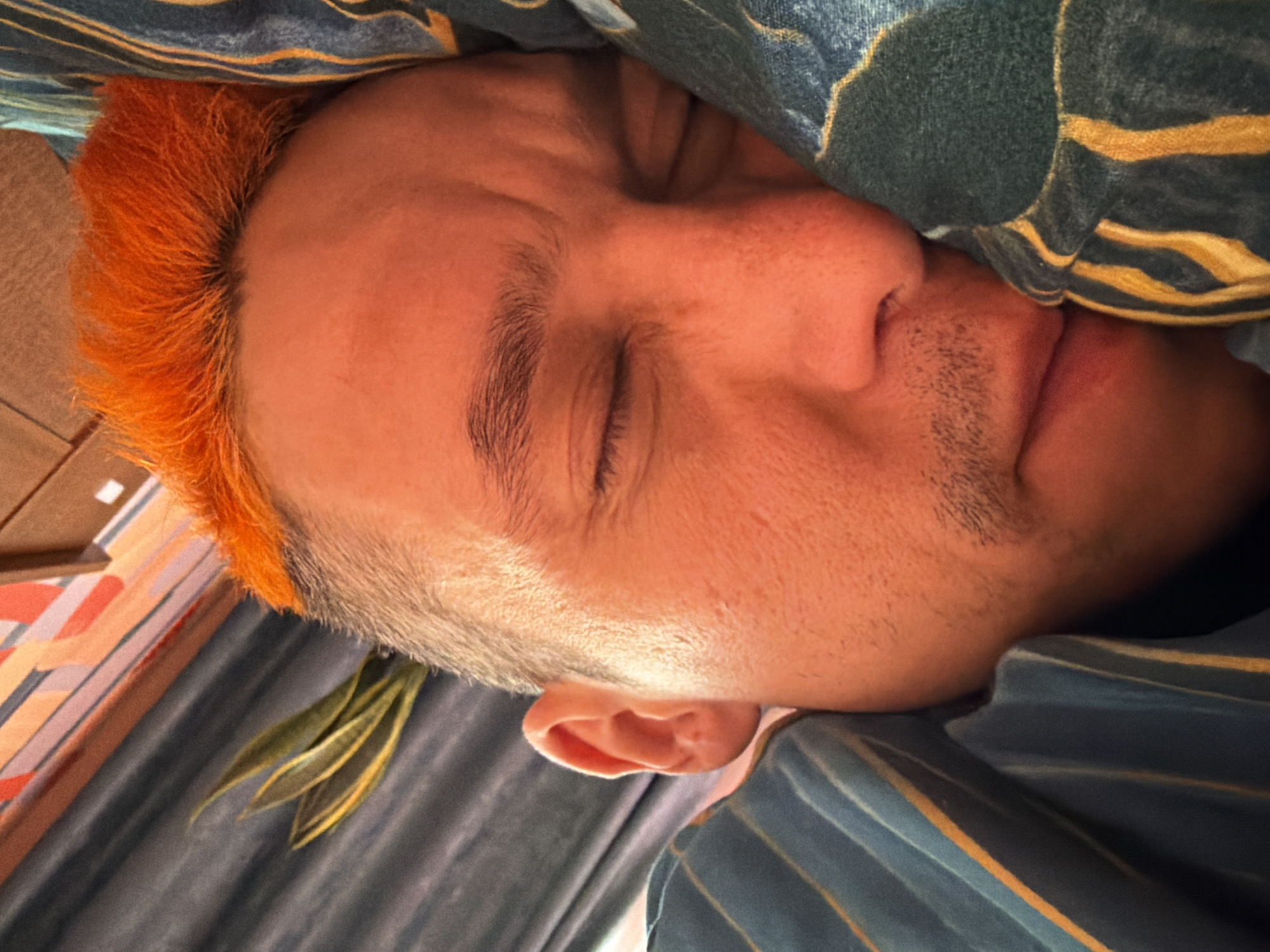 A close-up, indoor photograph shows a person sleeping. The person has orange hair on the left side of the frame and is partially covered by bedding. The person's eyes are closed, and the mouth is slightly open. The bedding appears to be a combination of patterned and striped fabric. The location is unspecified and not detectable from the image. The city of Chaikovsky, Russia is not detectable from the image.
