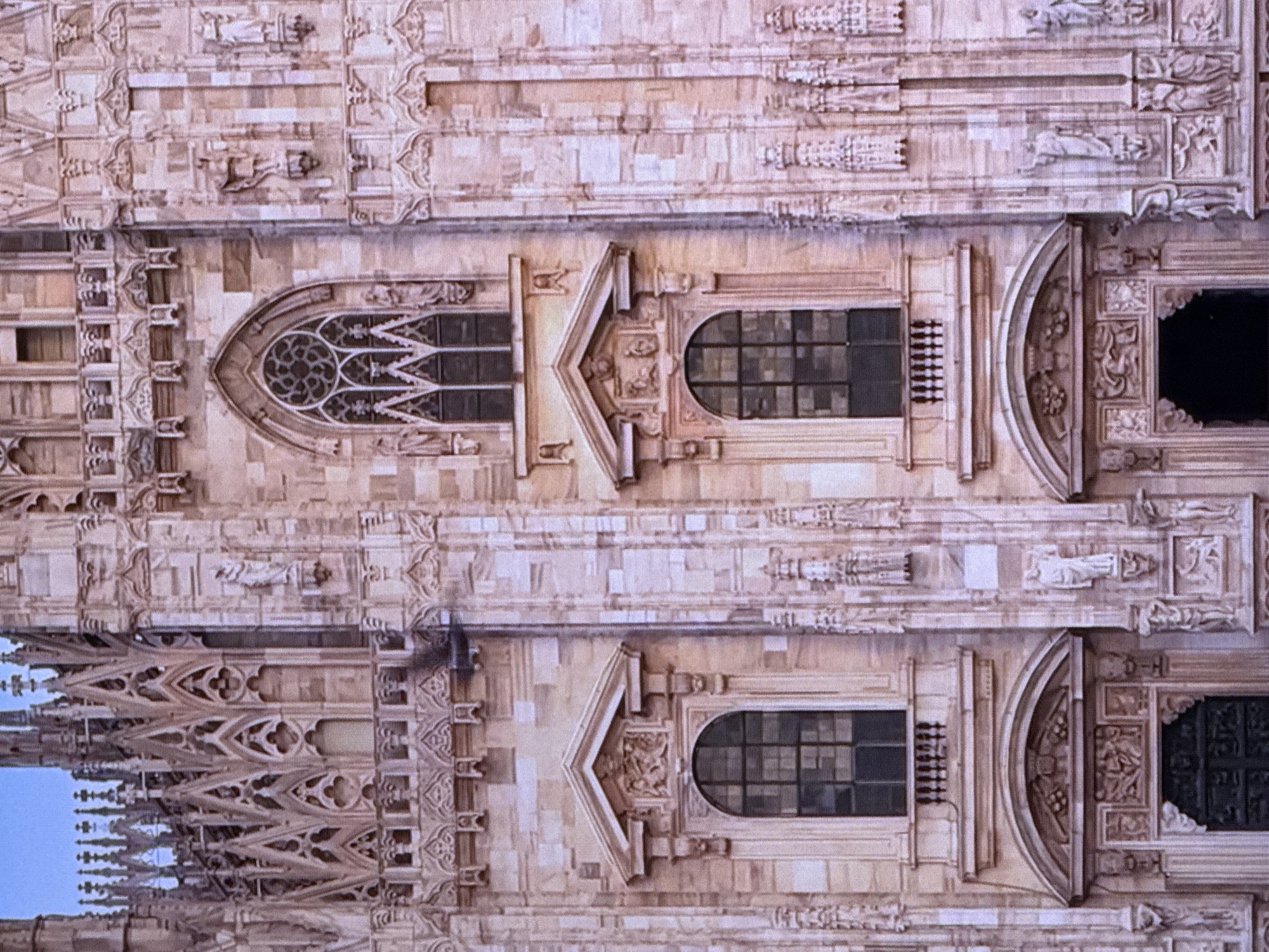 The image shows a close-up view of the intricate facade of a Gothic cathedral, characterized by its detailed stonework, pointed arches, and decorative carvings. Several ornate windows are visible, including a large rose window at the top and smaller arched windows below. Statues of figures adorn the facade, integrated into the architectural design. The stone appears to be a light-colored sandstone, weathered with age. The sky is a clear blue. The image does not depict any elements that would place it in Dubai, United Arab Emirates; it is consistent with European Gothic architecture, such as Milan Cathedral. There are no people or actions depicted.