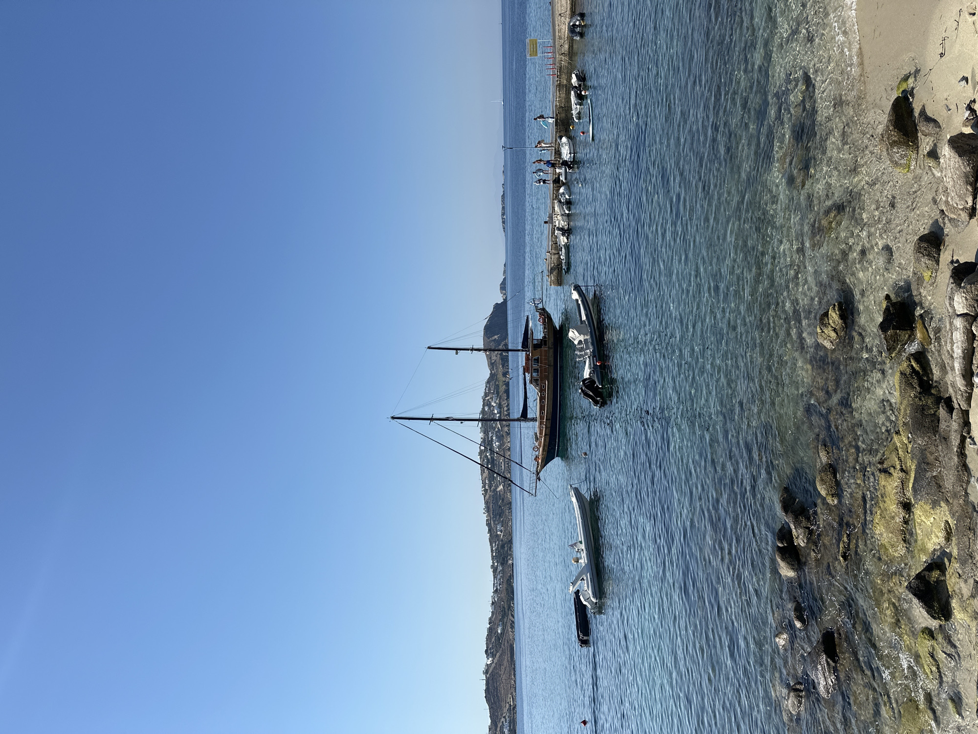 A wide-angle, eye-level, vertical shot captures a bright, sunny day at a bay, likely in Agios Ioannis Diakoftis, Greece, with clear blue sky dominating the upper portion of the image. The water is calm and transparent, revealing a rocky seabed near the shore and gradually becoming deeper blue further out.
In the foreground, a sandy beach meets the water's edge, littered with various sizes of rocks, some covered in green algae. The clear water laps gently against the shore.
Further into the bay, several small inflatable boats with outboard motors are visible. One white inflatable is prominent in the mid-left of the frame, with a person sitting inside. Another dark inflatable is partially submerged to its left.
Dominating the mid-ground is a large, traditional wooden sailboat with two masts, anchored in the bay. Its hull is dark blue, and the upper structure is wooden.
To the right of the sailboat, a concrete pier extends into the water. Several small boats are docked alongside it. A group of approximately seven people, dressed in casual, light-colored clothing, are standing on the pier. Some appear to be interacting or observing the boats. A yellow sign is attached to a post at the end of the pier.
In the background, a hillside dotted with white buildings stretches along the coastline, indicative of a settlement. The distant horizon shows the sea meeting the sky under the clear, bright conditions. The overall scene depicts a tranquil coastal environment with recreational boating activities.