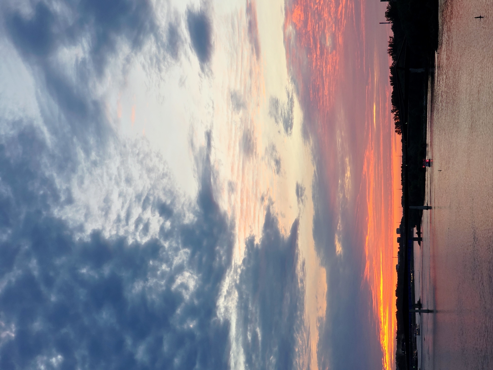 The image depicts a sunset over a body of water, likely a river, in Warsaw, Poland. The sky is a dramatic canvas of colors, with vibrant oranges and pinks bleeding into softer yellows and blues. Wispy clouds are scattered across the sky, some dark and moody, others catching the golden light of the setting sun.
Along the far bank, a dense line of trees forms a dark silhouette against the colorful sky. Several structures are visible through the trees, including what appears to be a bridge with its supports extending into the water. On the water itself, there are a few small boats, one with its lights on, and a few poles marking the edge of a channel or dock. The water reflects the colors of the sky, creating a shimmering, ethereal effect.
The time of day is clearly sunset, indicated by the intense colors in the sky and the long shadows that would be cast. The weather appears calm, with no strong winds suggested by the smooth water and the way the clouds are positioned. The overall emotion evoked by the image is one of peacefulness and awe at the beauty of nature. There is no visible text in the image.