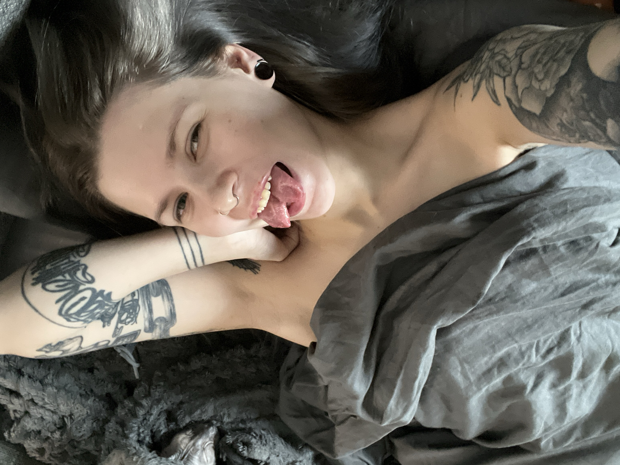 A person is lying in bed, partially covered by a gray sheet or blanket. They have dark hair, light skin, and visible tattoos on their arms. The person is sticking their tongue out and looking towards the camera. The environment appears to be a bedroom setting, with a darker texture possibly being a part of the bed or bedding at the bottom of the frame. There is no indication of the image being taken in the city of Khimki, Russia.