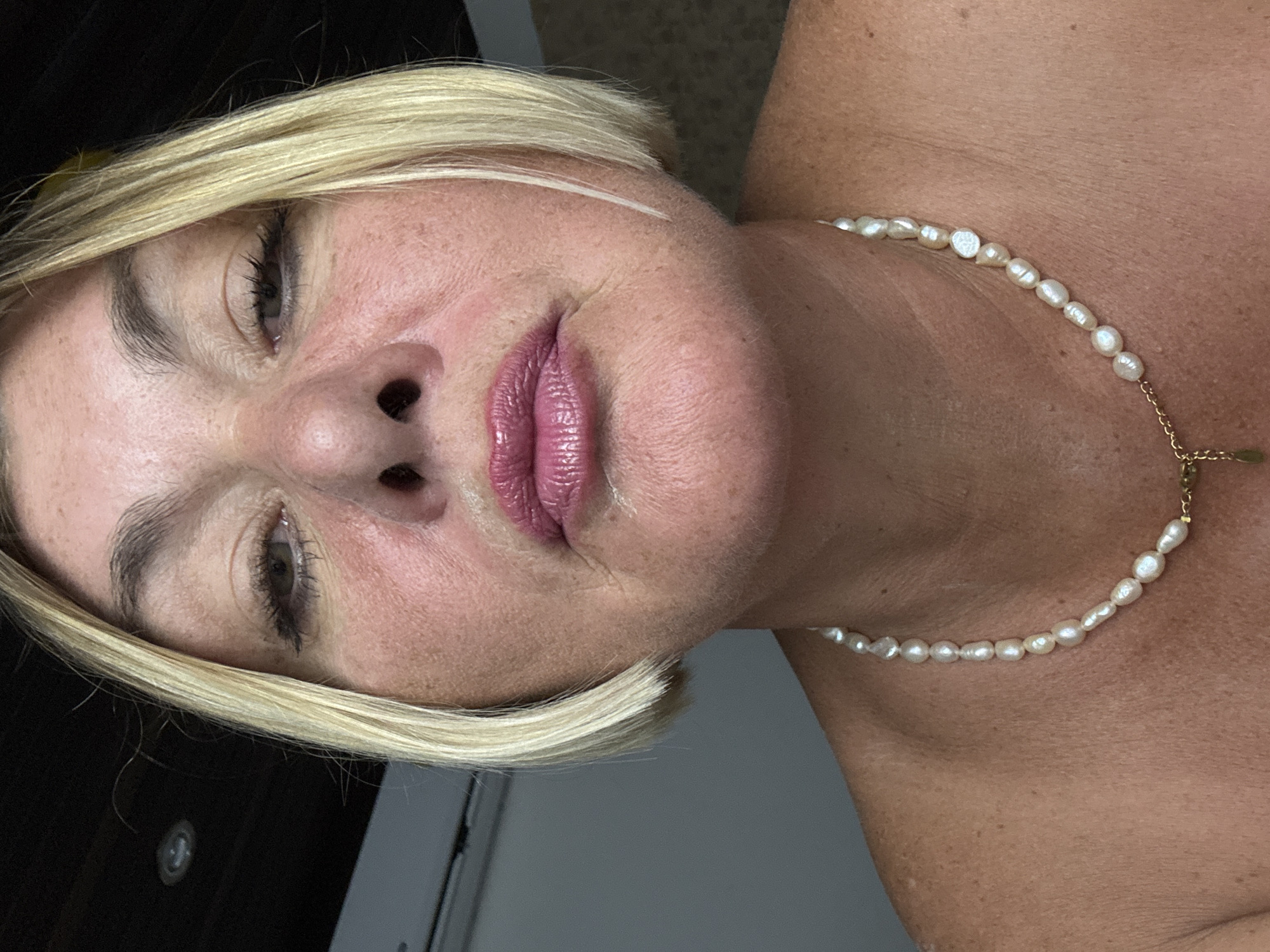 The image is a close-up portrait of a fair-skinned woman with blonde hair. She is wearing a pearl necklace. The location is not evident.