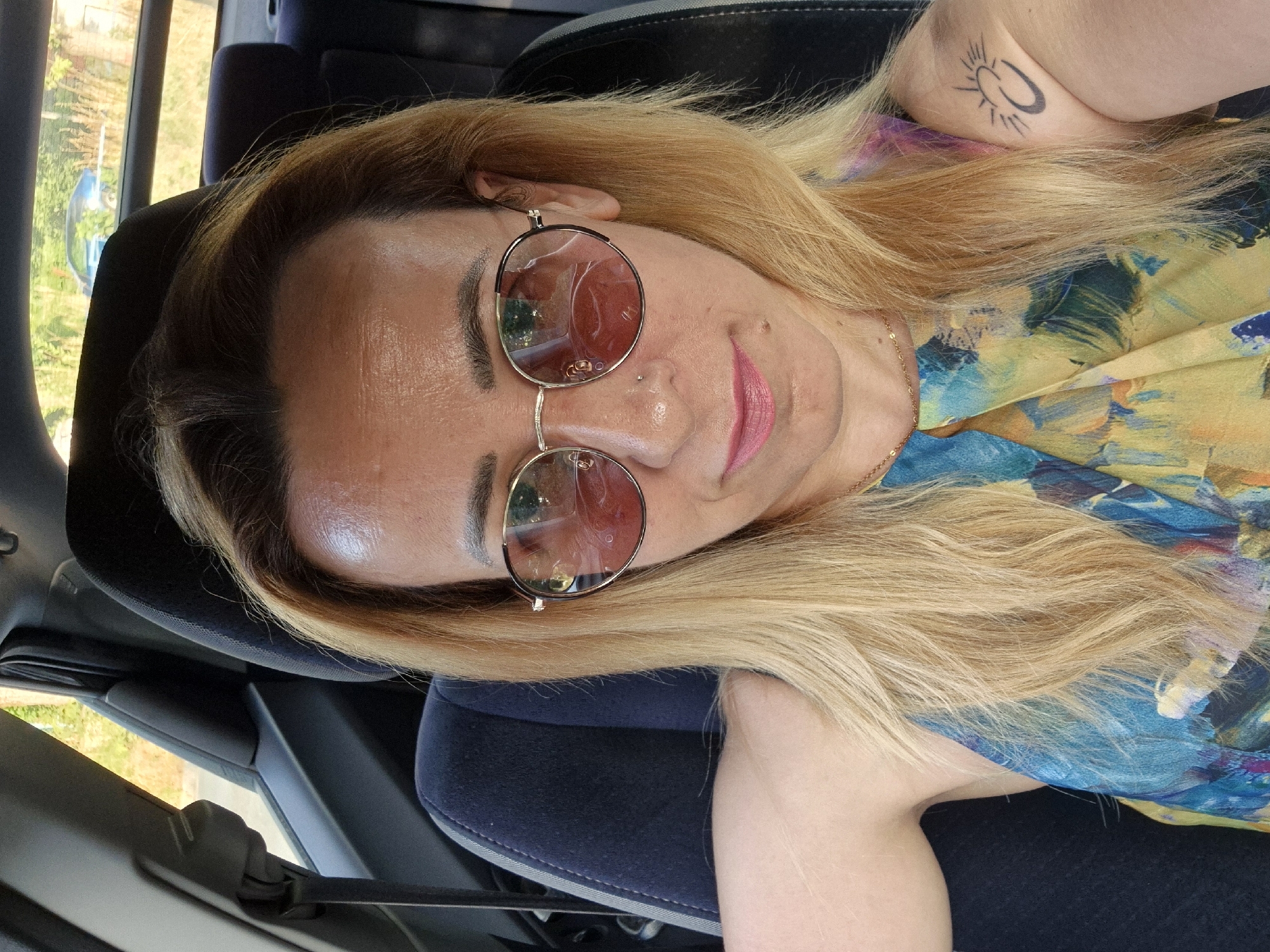 The media file is a selfie taken by a person inside a vehicle. The person has blonde hair and is wearing round, pink-tinted sunglasses. They have on a colorful, patterned top and a thin gold necklace. A tattoo of a sun with a crescent moon is visible on their upper arm. The person is smiling slightly and looking towards the camera. The setting appears to be daytime, with sunlight visible through the car window, suggesting a clear day. The interior of the car is dimly lit, with dark-colored seats. The background outside the window shows greenery and trees, indicating an outdoor location, possibly in a rural or suburban area. The overall mood conveyed by the image is casual and relaxed.