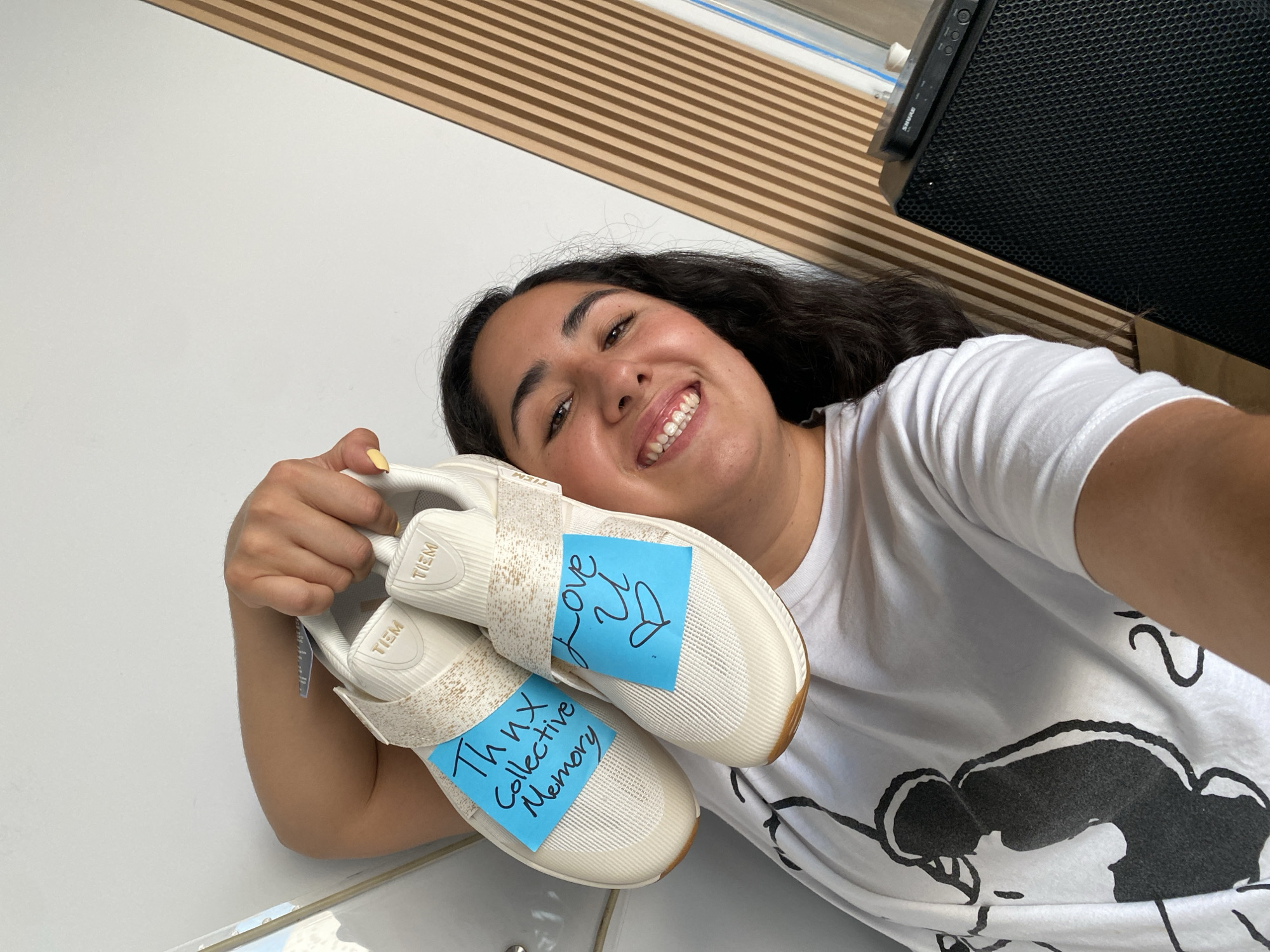 A young woman with dark, curly hair and a wide, happy smile is captured in a bright, indoor selfie, likely taken in Puebla, Mexico. She is lying down, looking directly at the camera, exuding a cheerful and relaxed demeanor.
She wears a white short-sleeved t-shirt with a large, stylized black graphic partially visible on the chest. Her fingernails are painted a light yellow. She is holding up a pair of cream-colored athletic shoes, possibly designed for studio or fitness activities, which feature a knit-like texture, light brown soles, and a Velcro strap. The brand name "TIEM" is visible on the heel of both shoes.
Attached to the shoes are two light blue sticky notes with handwritten messages. One note reads "Thnx Collective Memory," and the other says "Love U ❤️" (with a heart symbol).
The background features a plain white wall on the left, and a section of light brown wooden slat paneling extends diagonally across the top right of the frame. Part of a black audio speaker or equipment, bearing the "SHURE" logo, is visible in the upper right corner, resting near the wooden slats. The bright lighting suggests it is daytime.
