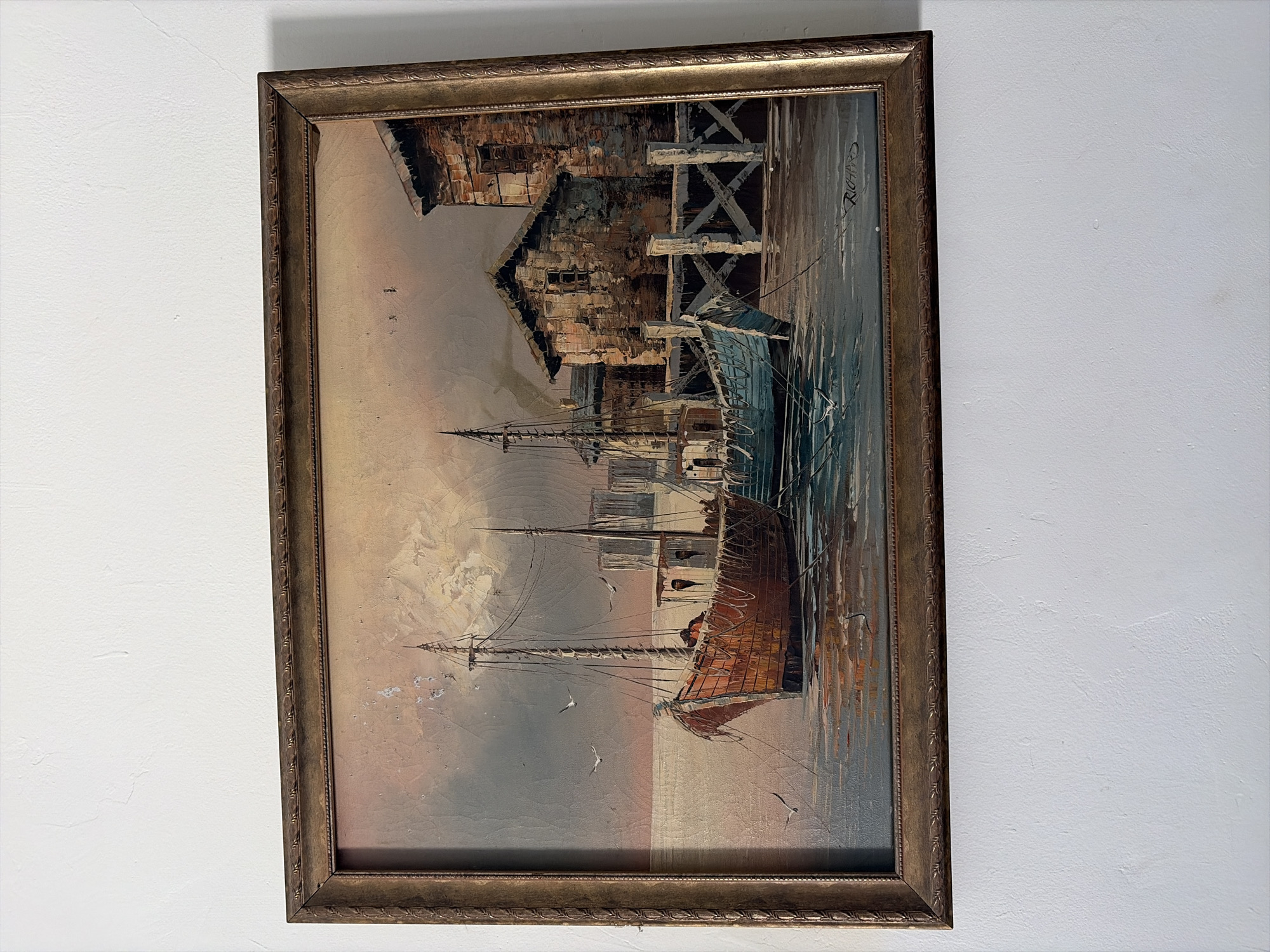 A framed painting hangs on a white wall. The painting depicts a harbor scene with boats, buildings, and a grey sky. The buildings are detailed with visible brushstrokes and seem to be old, possibly representing a historical waterfront. The frame is ornate and metallic, with a dark brown or bronze color. The painting is signed 'RICHARD' in the upper right corner.